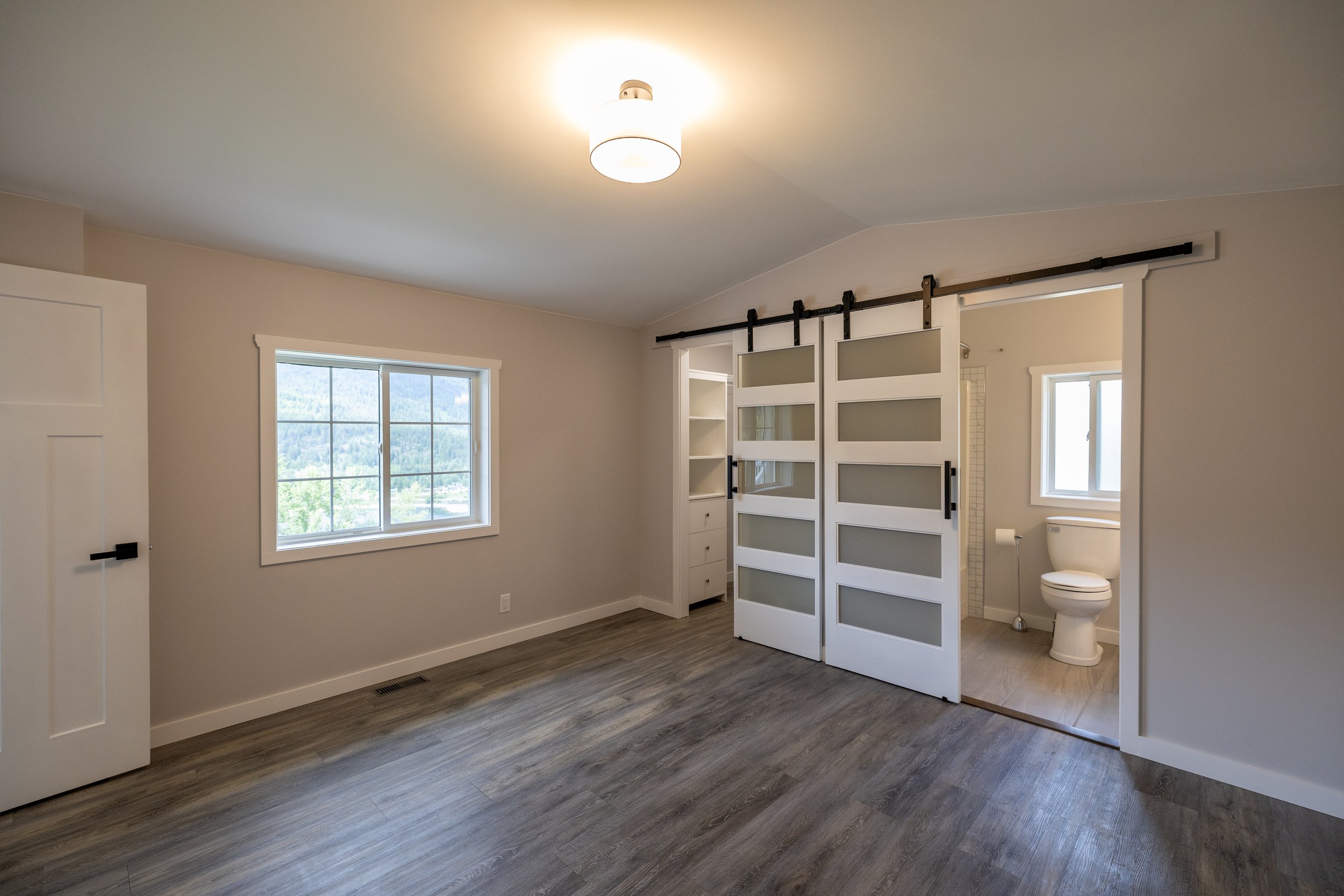 Empty bedroom with wood flooring, white walls, two windows, a ceiling light, and a sliding barn door leading to a bathroom with a toilet and window.