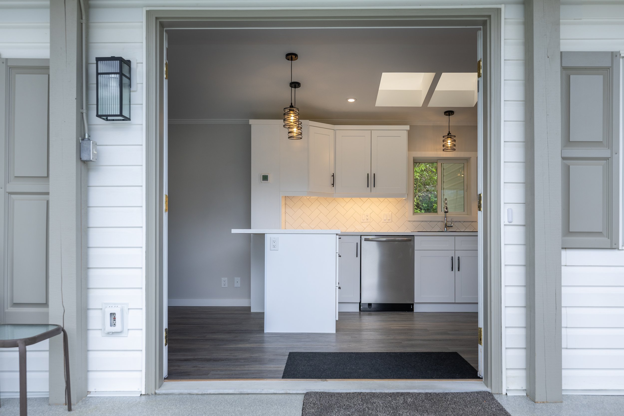 View of a modern kitchen seen through a large open doorway, with white cabinets, a small window, stainless steel dishwasher, pendant lighting, and hardwood flooring.