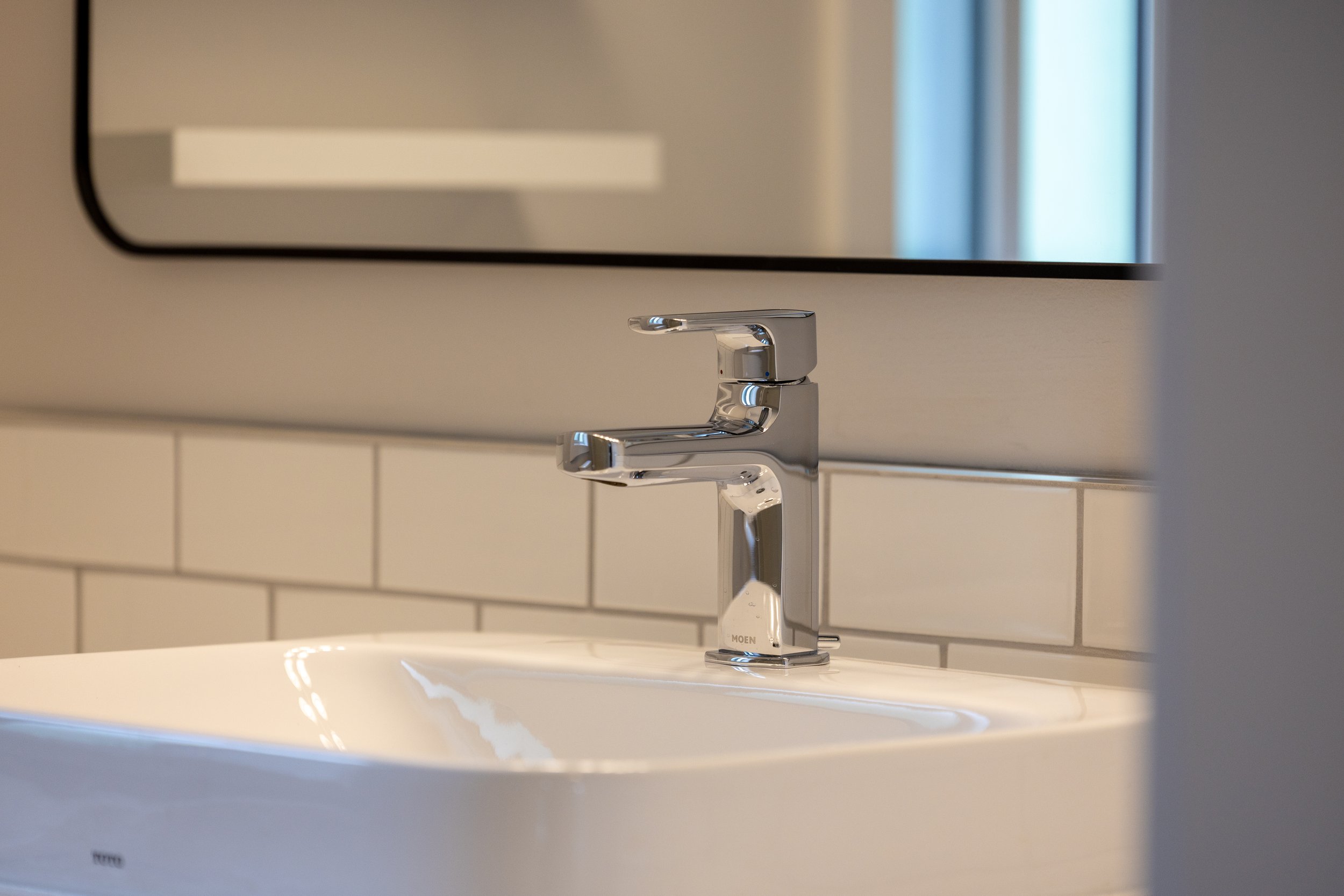 Close-up of a modern chrome bathroom faucet above a white sink with beige tiled wall and mirror reflection in background.