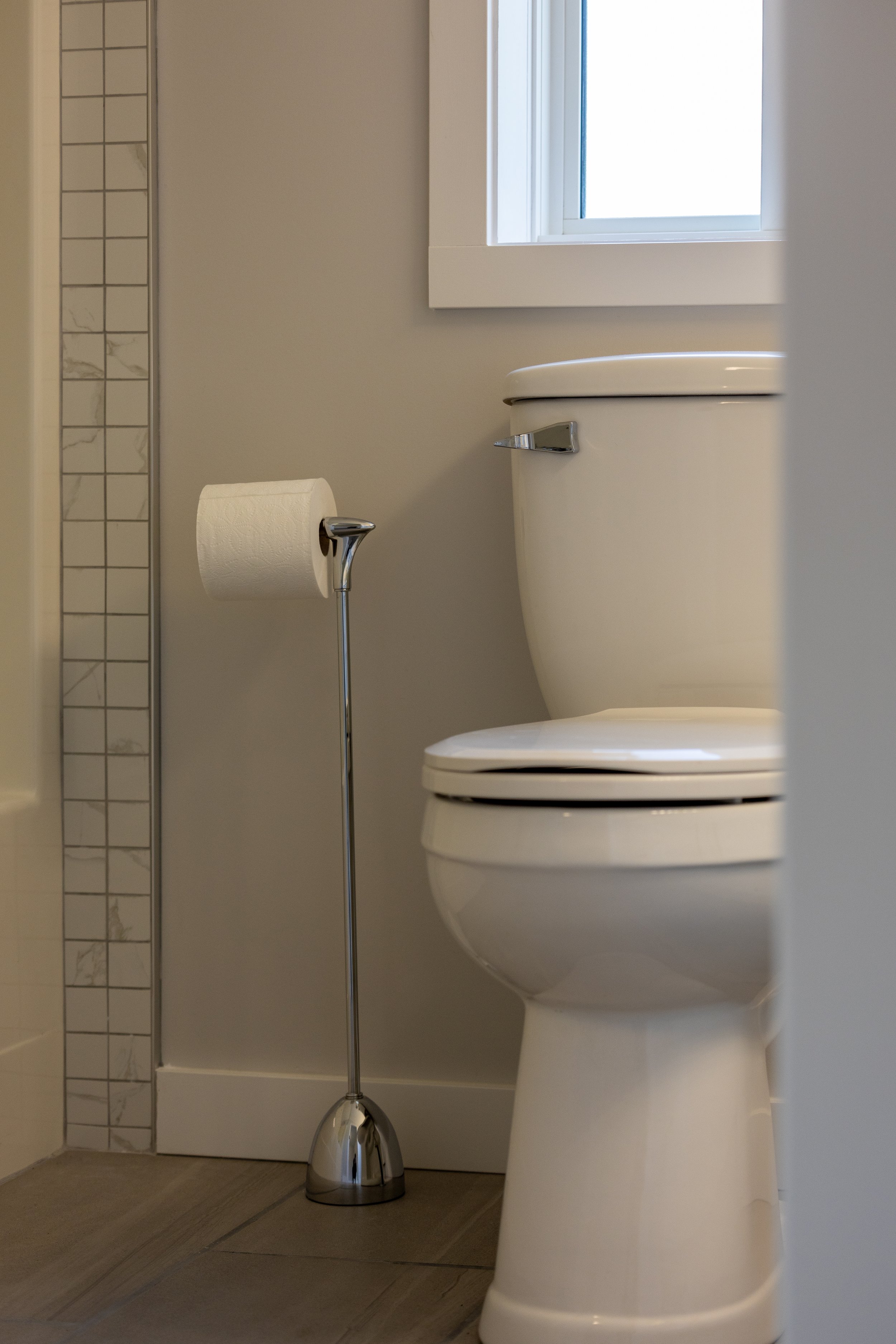 Close-up of a white toilet next to a window, with a chrome toilet paper holder on a stand nearby in a bathroom.