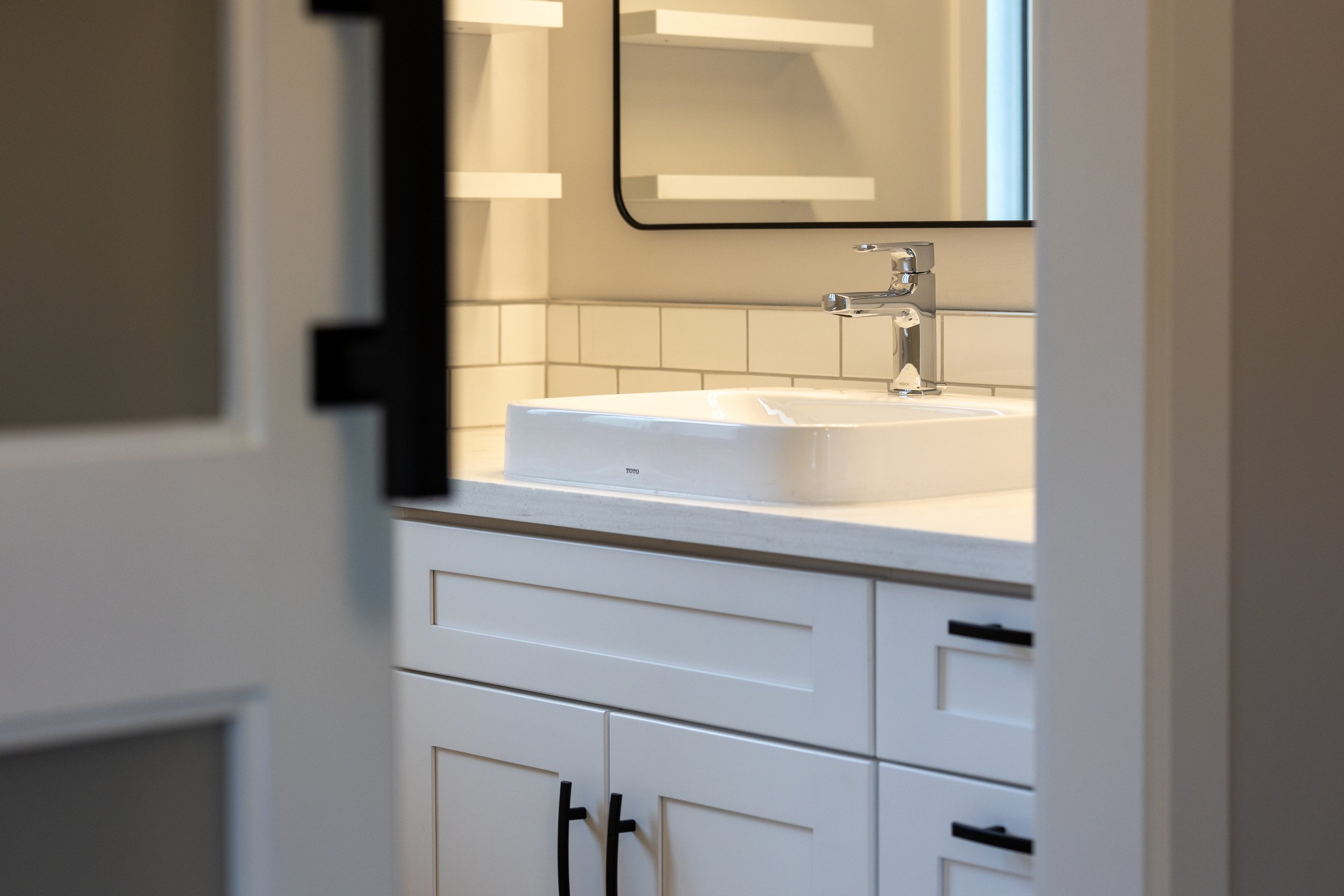 Bathroom with a white sink, black cabinet handles, mirror, white tile backsplash, and white cabinet with black handles