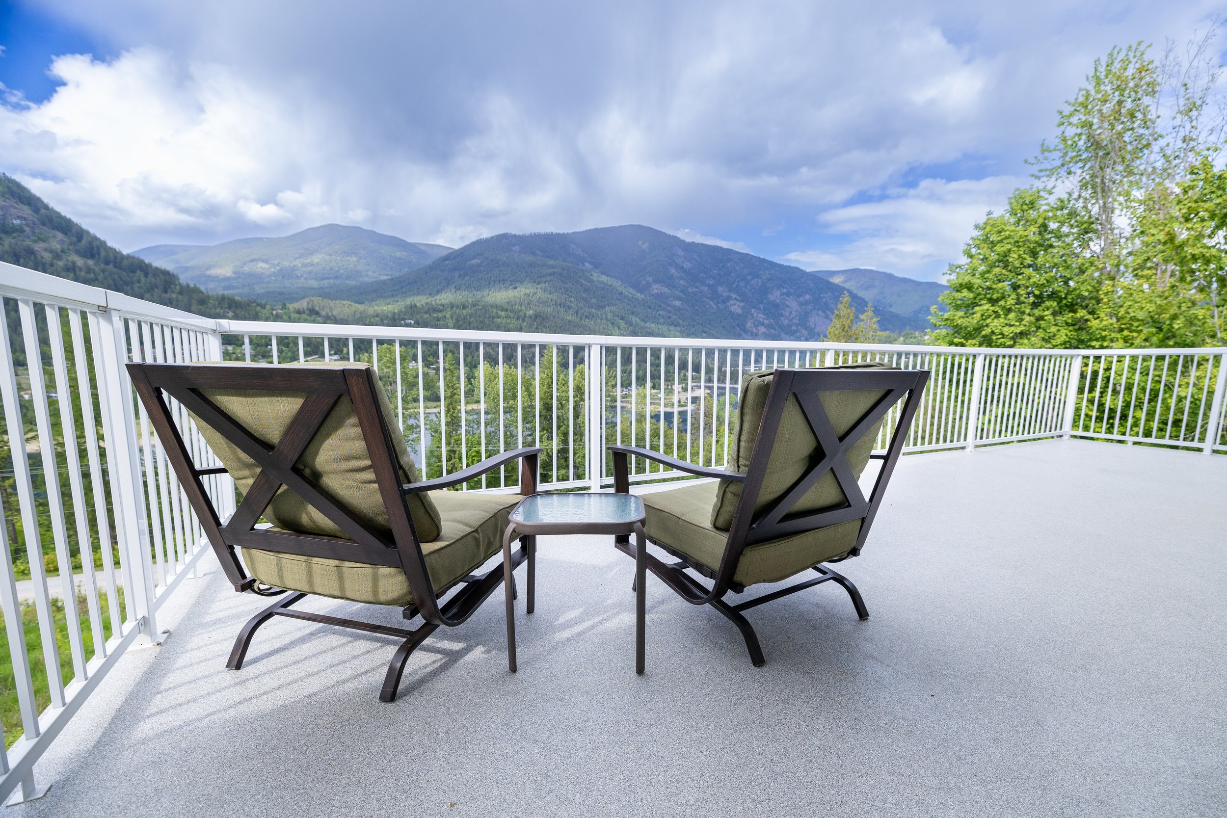 Two cushioned outdoor chairs with a small table between them on a balcony overlooking a scenic mountain view with green trees and cloudy sky.