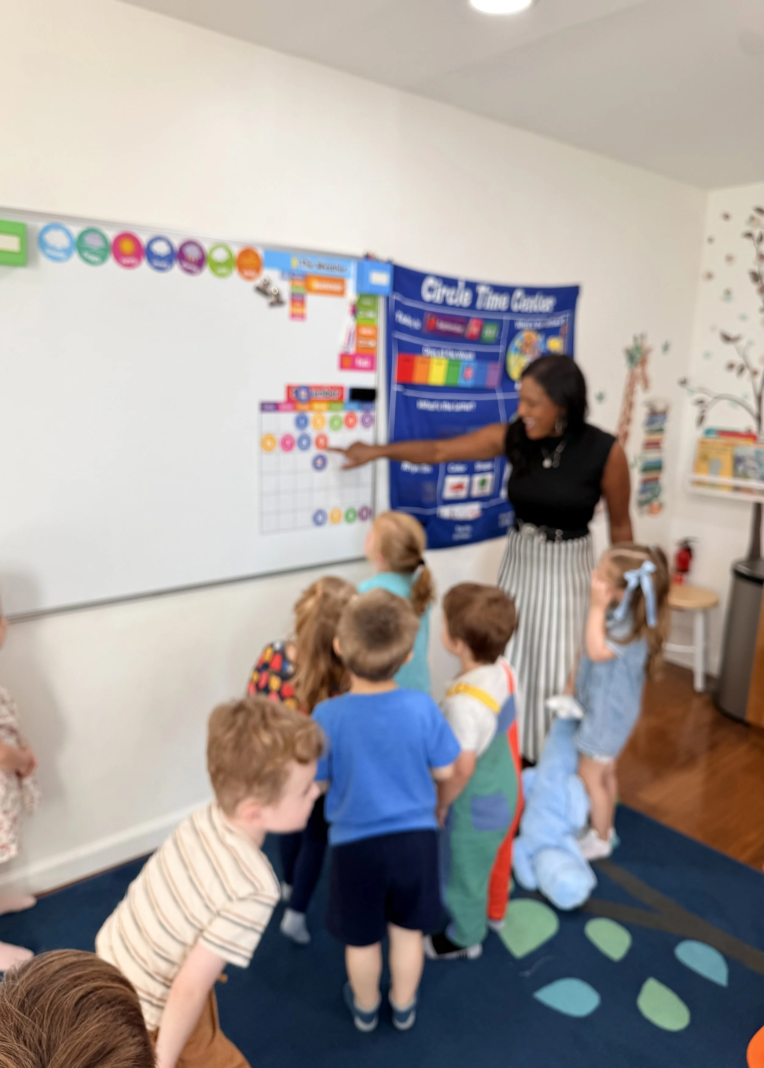 Two preschoolers plaMiss Natasha leading morning meeting with preschoolers at the calendar boardying side by side in the Discovery classroom