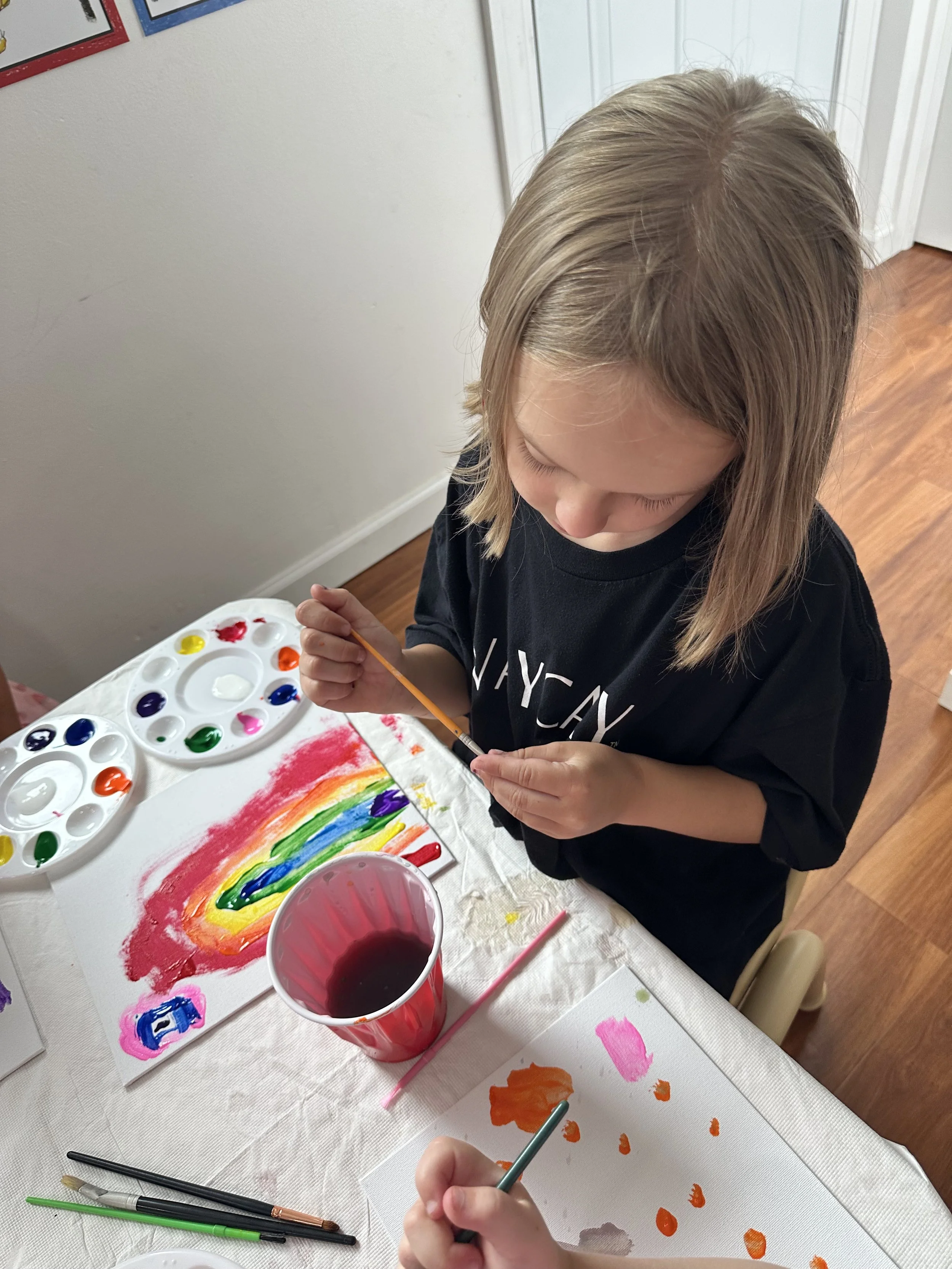 Two children smiling Preschooler painting a rainbow with watercolorsindoors near a fireplace, with one wearing a green sequined hat and green checkered dress.