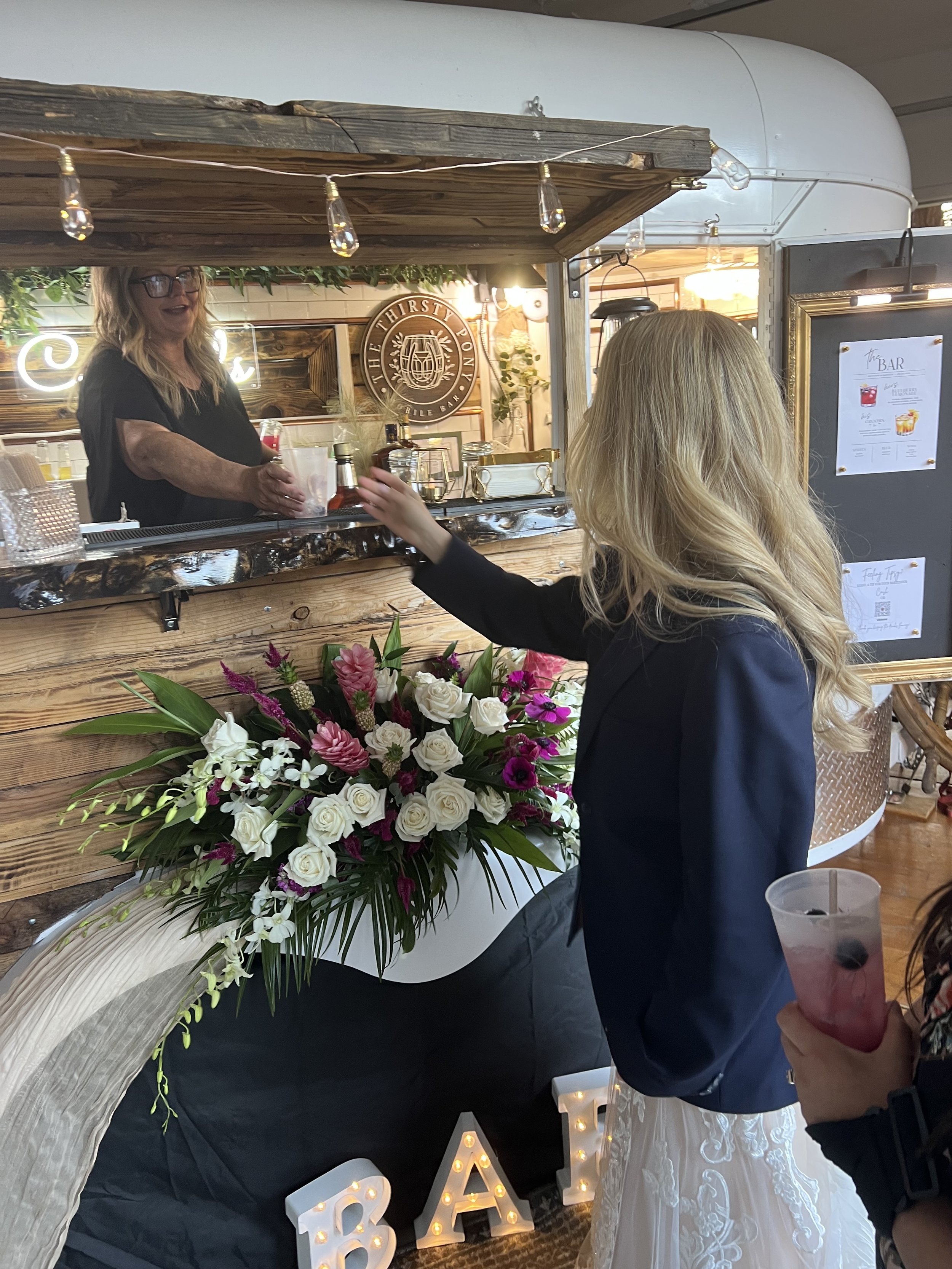 A bride is receiving a drink from a bartender at a wooden bar. There is a large floral arrangement with white and purple flowers next to her. The bar area has a rustic decor with string lights and flowers.