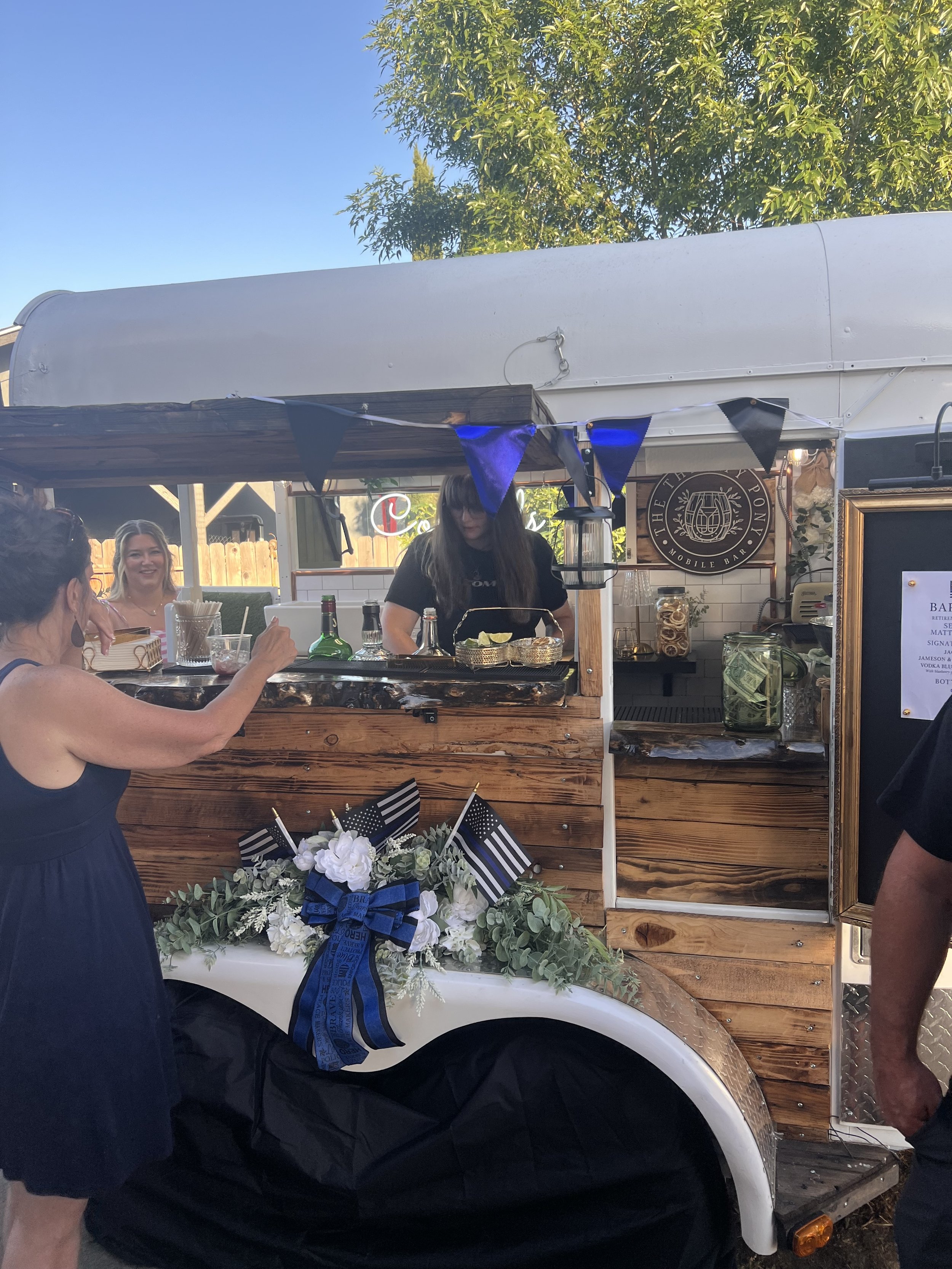 People order drinks at a vintage-style mobile bar trailer decorated with black, blue, and black flags, floral arrangements, and small flags of the United States and blue lines, with a bartender serving drinks and a woman in a black dress reaching out