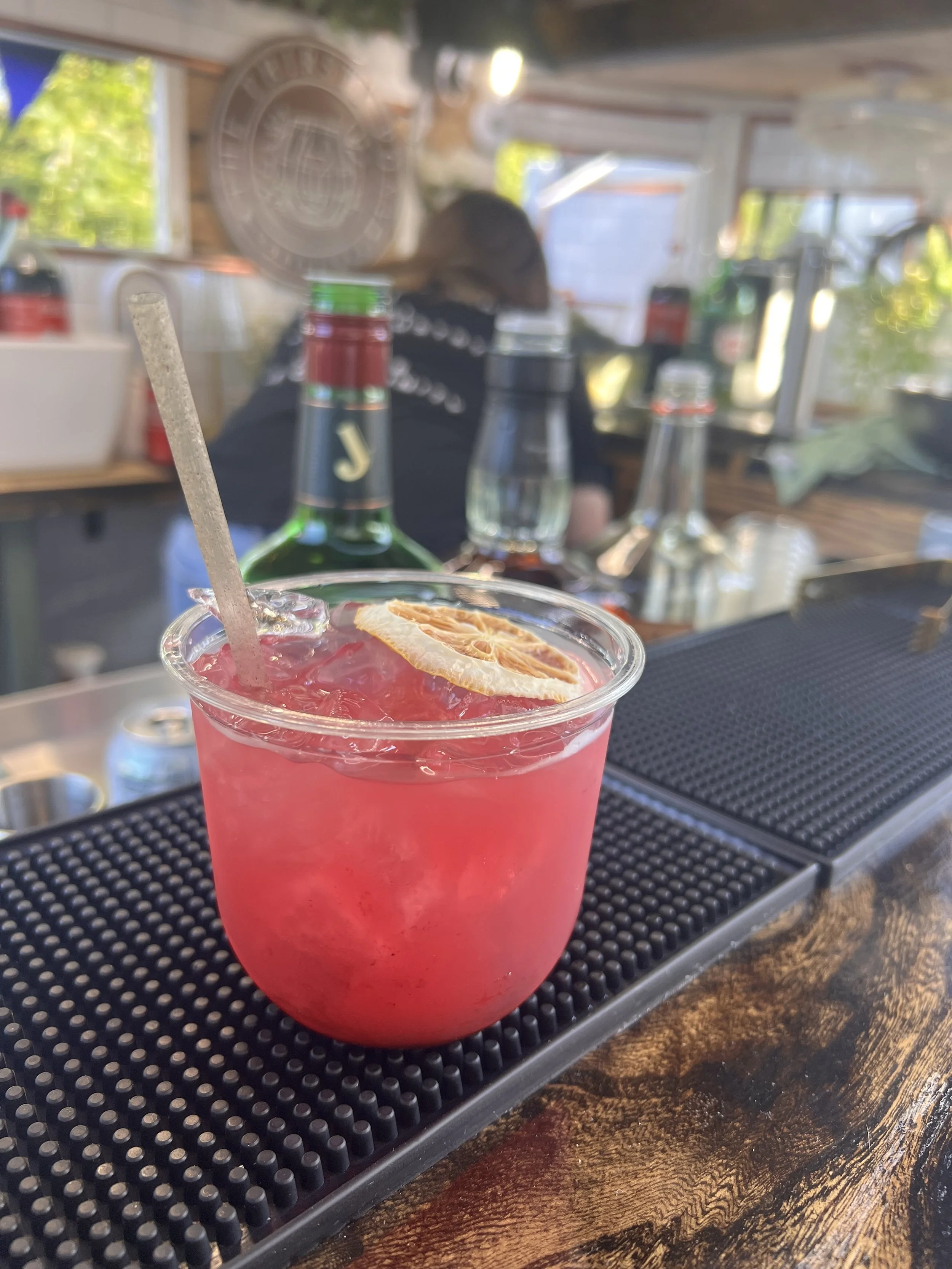 Pink cocktail with a lemon slice garnish and a straw on a bar counter, with bottles and a bartender in the background.