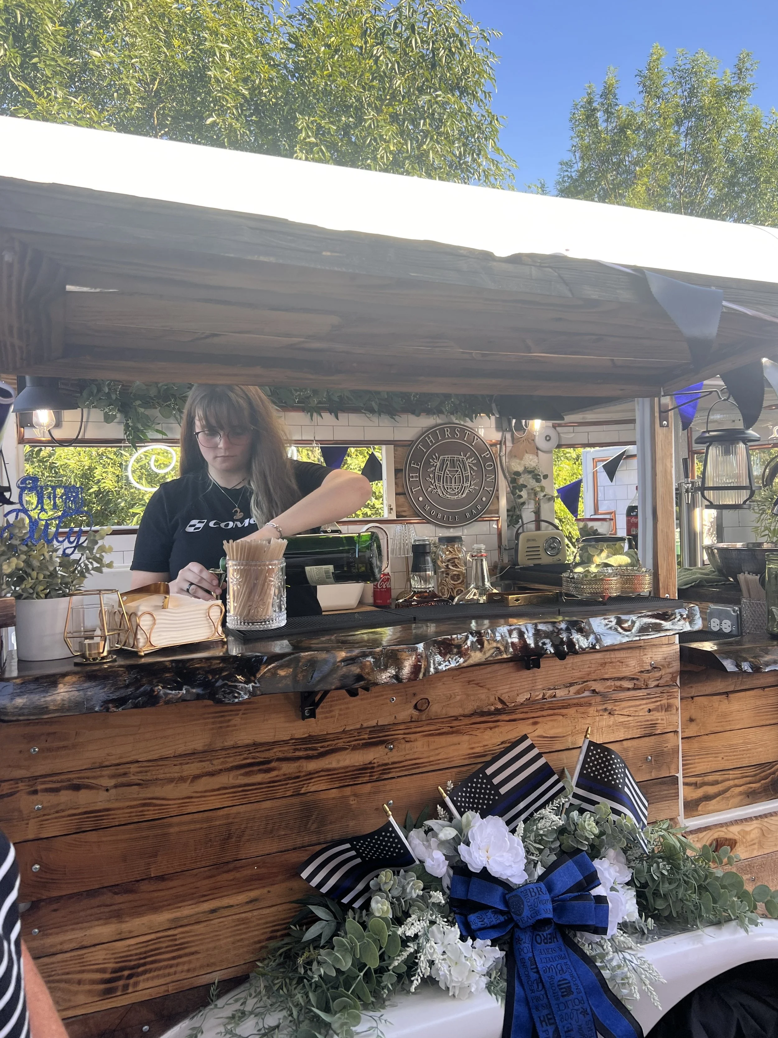 A woman bartending at an outdoor booth with a sign that reads 'The Thirsty Pony'. The booth has a wood countertop, various bottles, and bar tools. There are decorations including black and white American flags and a large floral wreath with white flowers, green foliage, and a blue ribbon. Bright sunlight and clear blue sky are visible above, with green trees in the background.