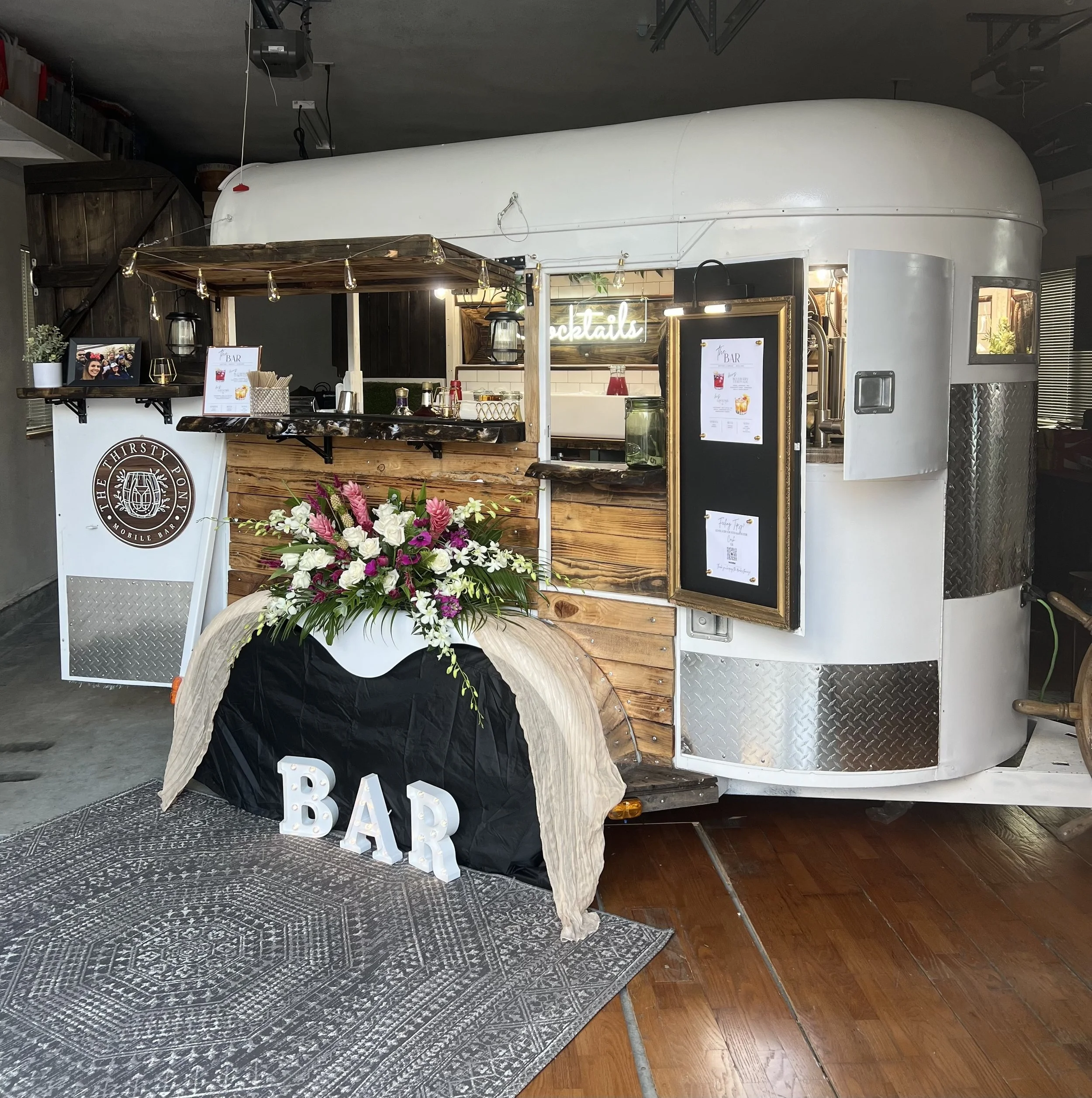 A vintage white trailer converted into a mobile bar with wood accents, floral decorations, and illuminated signs at a venue.