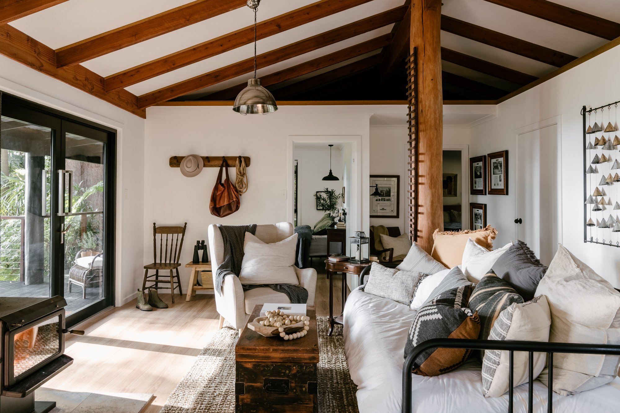 An interior shot of the cosy cabin with a tall wooden post supporting a wooden beamed roof. There is a white sofa with lots of brown, grey and grey cushions on one side of the image, and a deep white armchair in the centre of the image. The walls ar