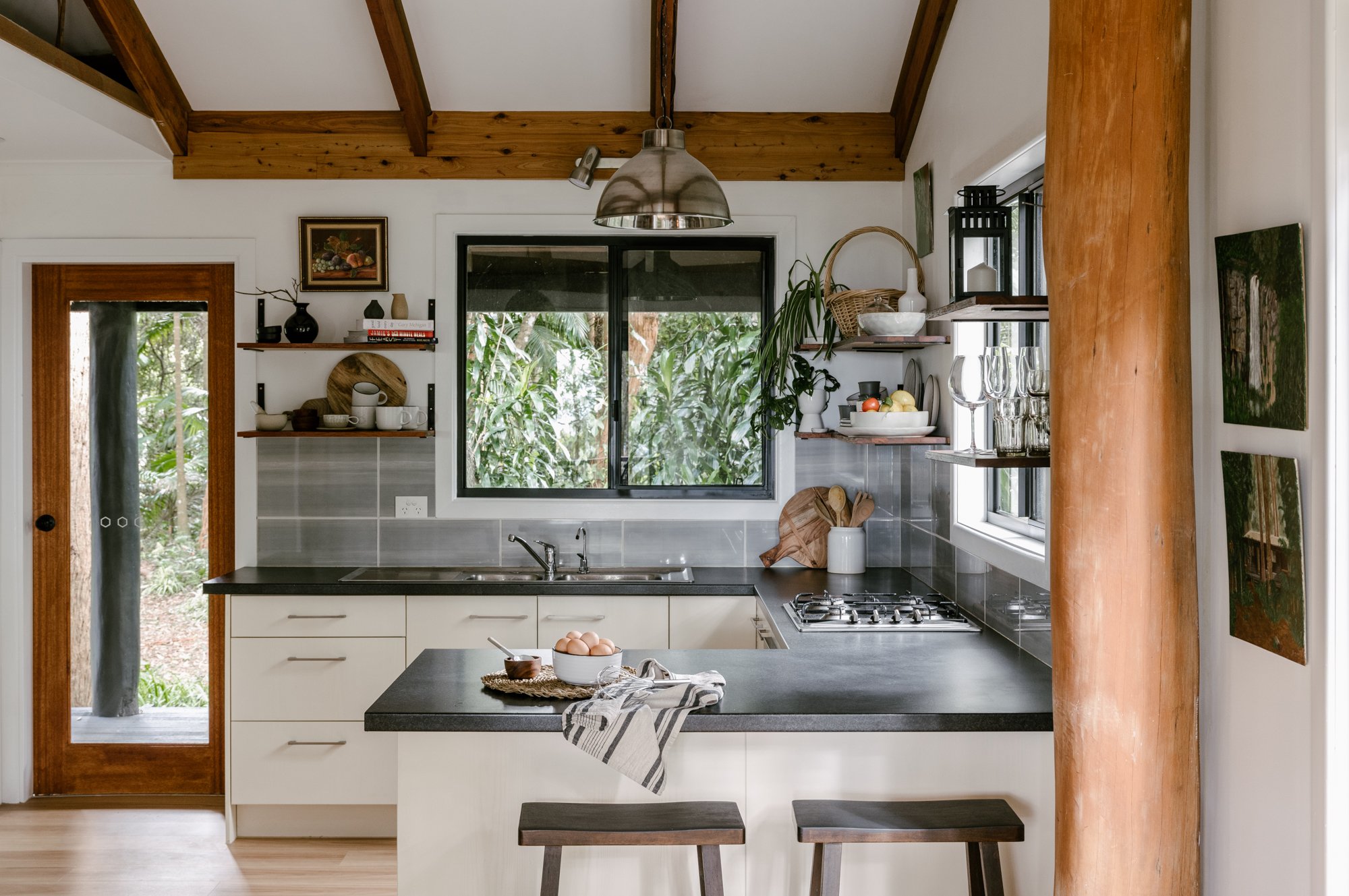 A picture of the kitchen with exposed wooden beams. The bench tops are dark grey and the under bench cupboards are cream. The splashback is grey. There are open shelves along two of the walls of the kitchen holding cups, vases, plates, plants and a