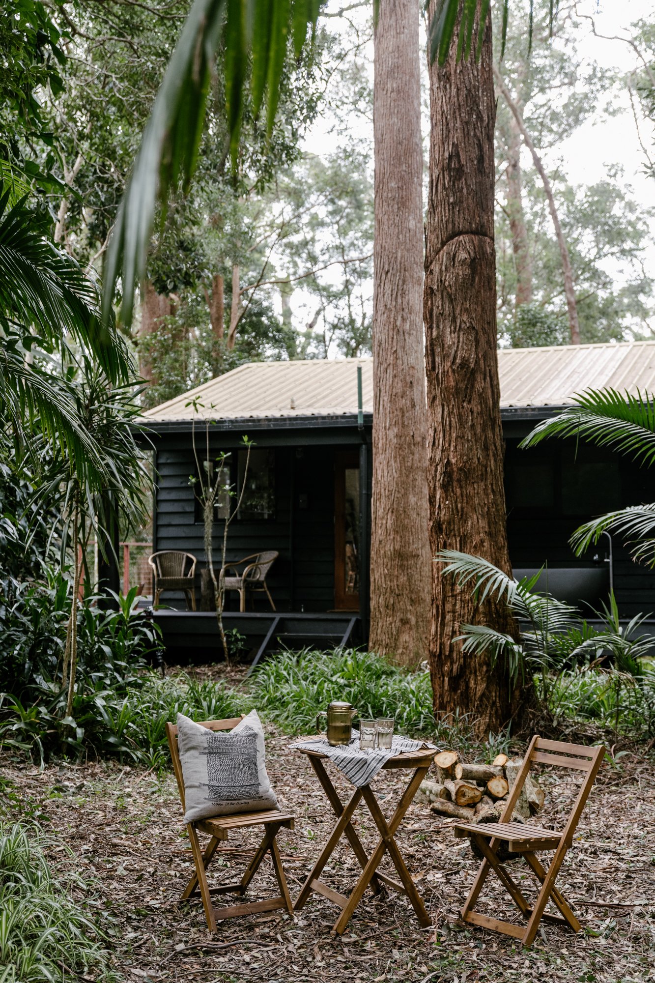 A back view of the black-painted cabin verandah. In the foreground are two chairs and a table in a clearing. There are two very large two trees in between this seating and the verandah. Through the trees, a glimpse of a back outdoor bath tub on the