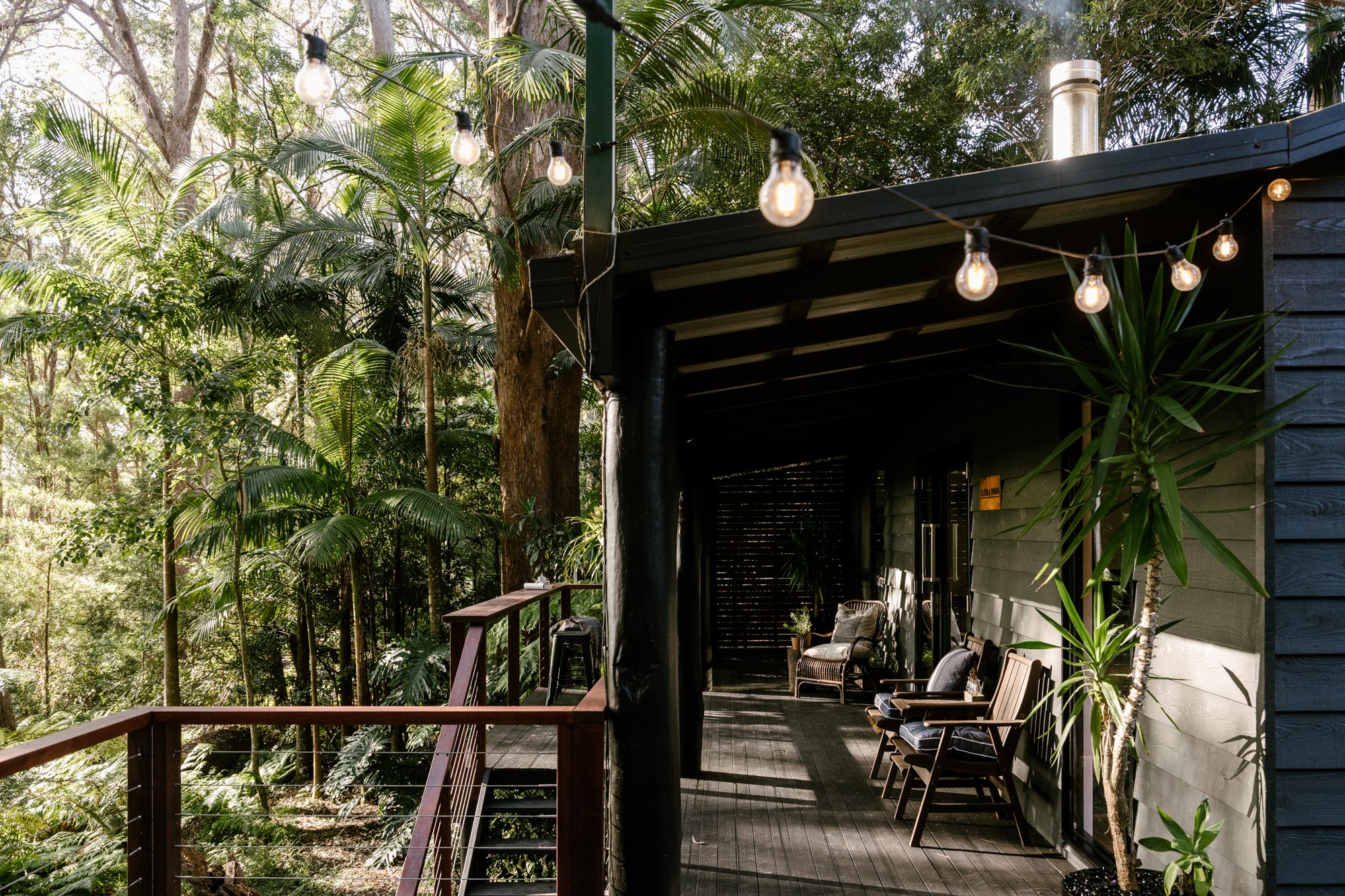 Front verandah of the black painted cabin with brown trim. There are comfortable and cushioned seats on the verandah and festive lights in the foreground of the image.