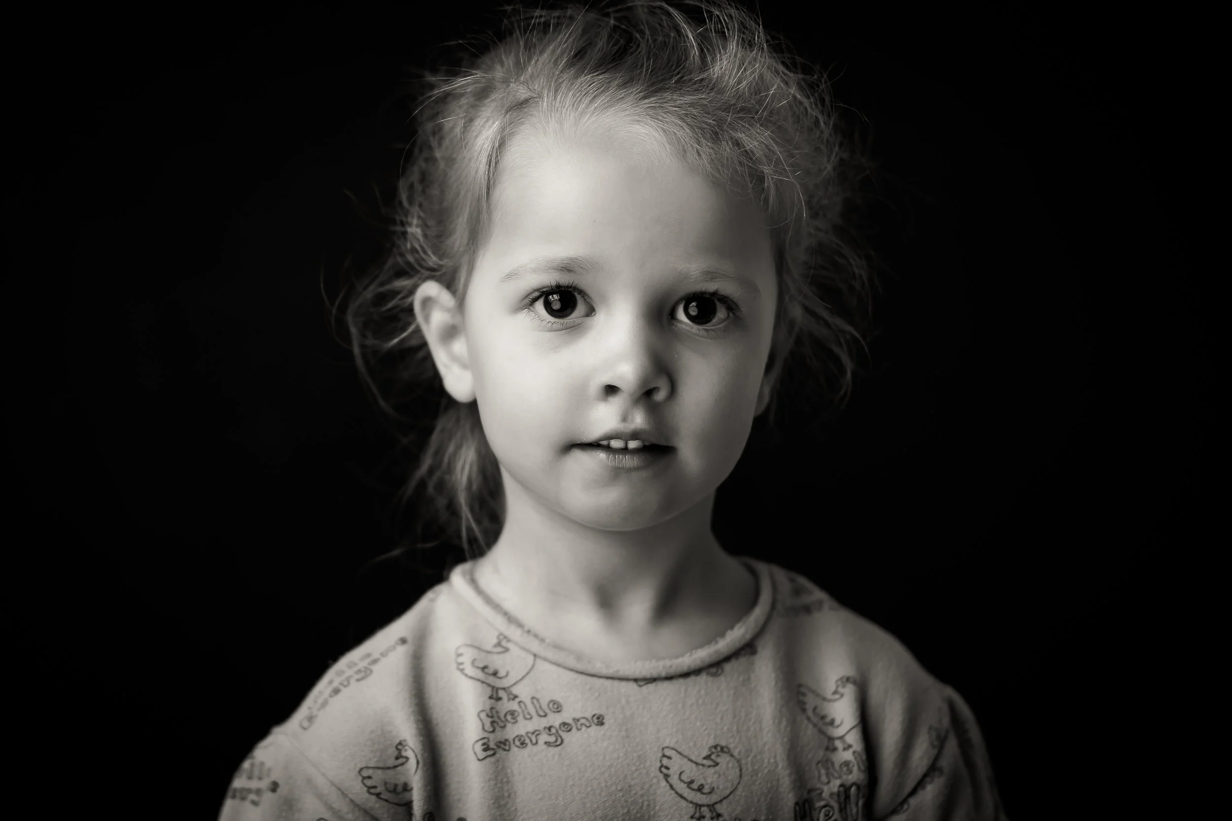 Black and white photograph of a young girl with wide eyes and slightly messy hair, wearing a shirt with a bird pattern. She is looking directly at the camera against a dark background.