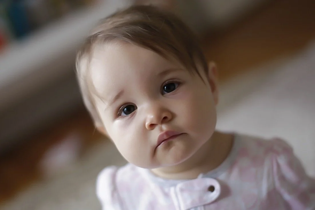 A young child with light brown hair and large dark eyes, wearing a light pink and white patterned shirt, looking at the camera with a neutral expression.