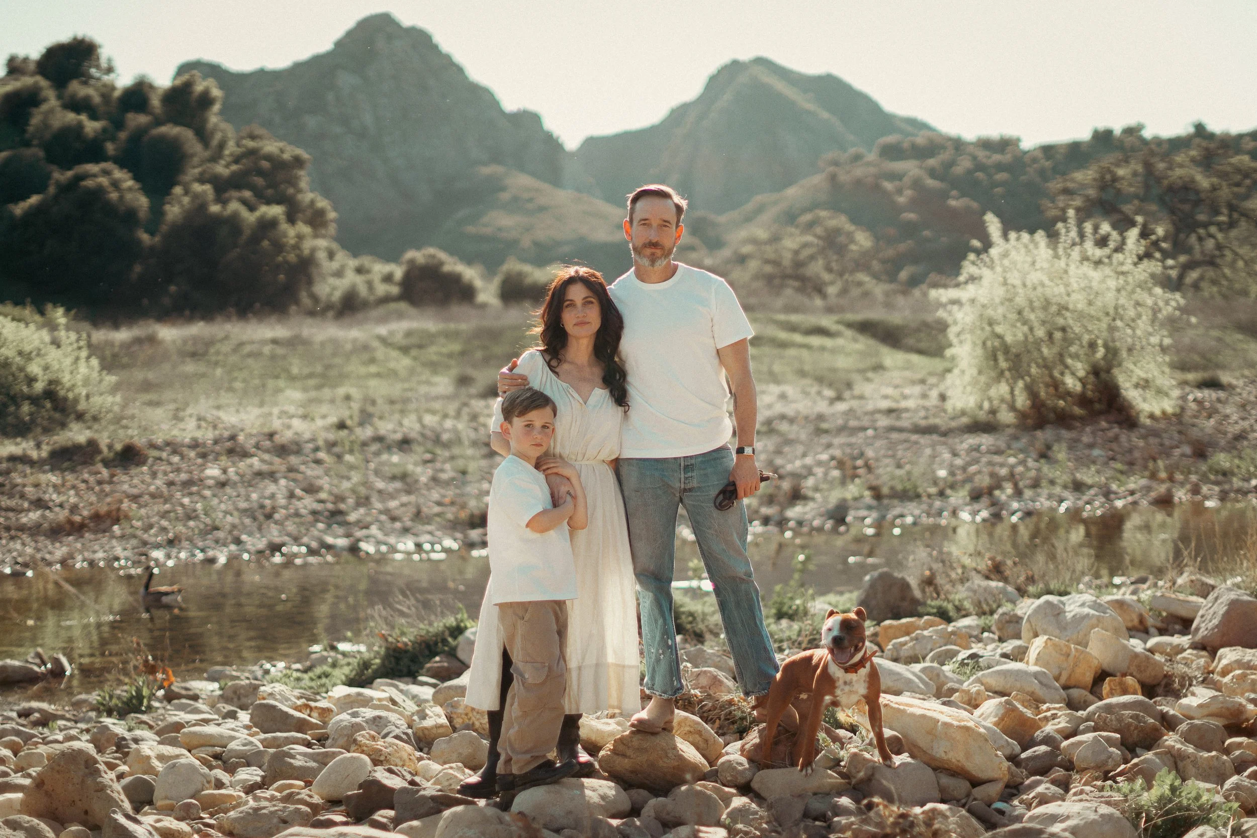 A family of three and their dog standing on a rocky riverbank with mountains in the background during daylight. The woman has dark curly hair and is wearing a white dress, standing in the middle with the man and child beside her. The man has a beard,