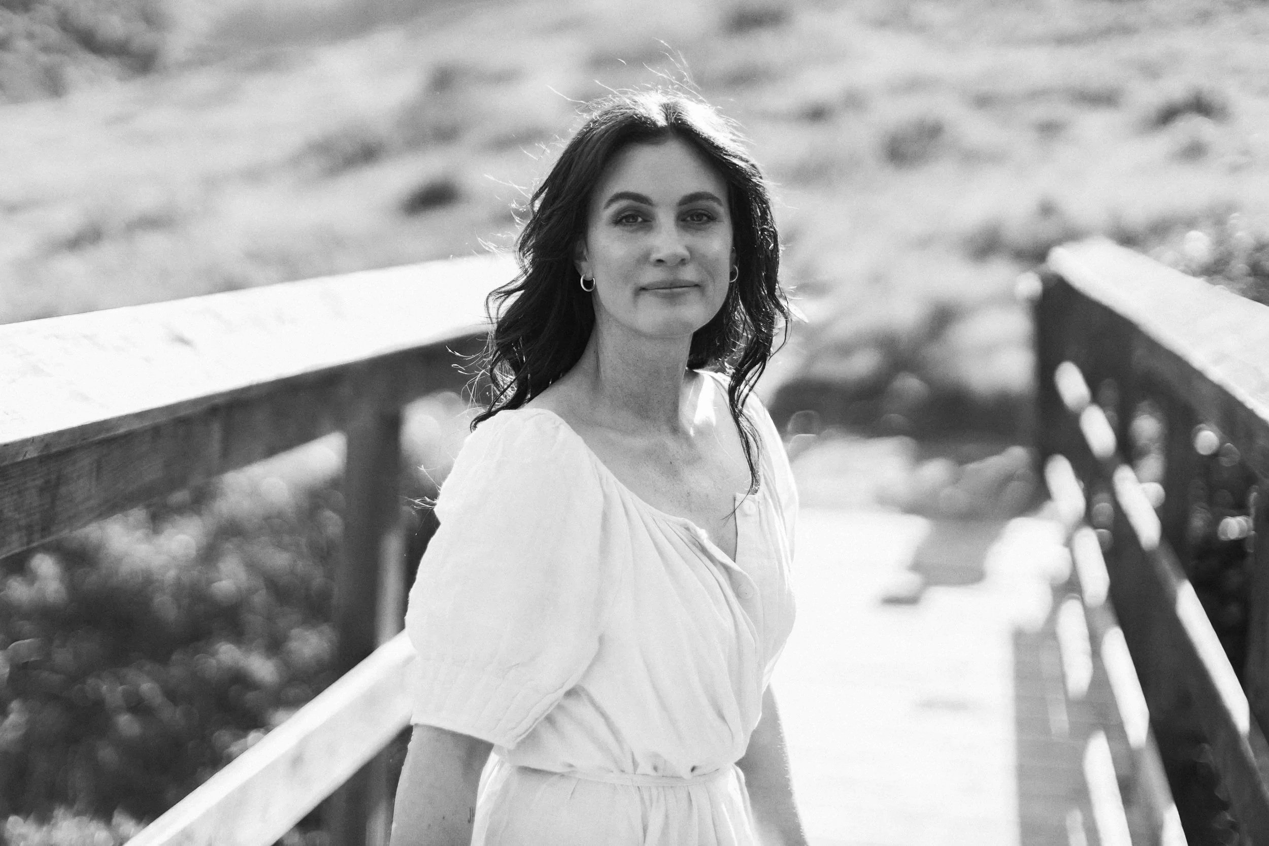 A woman with shoulder-length dark hair standing on a wooden bridge in a sunlit outdoor setting, wearing a light-colored dress.