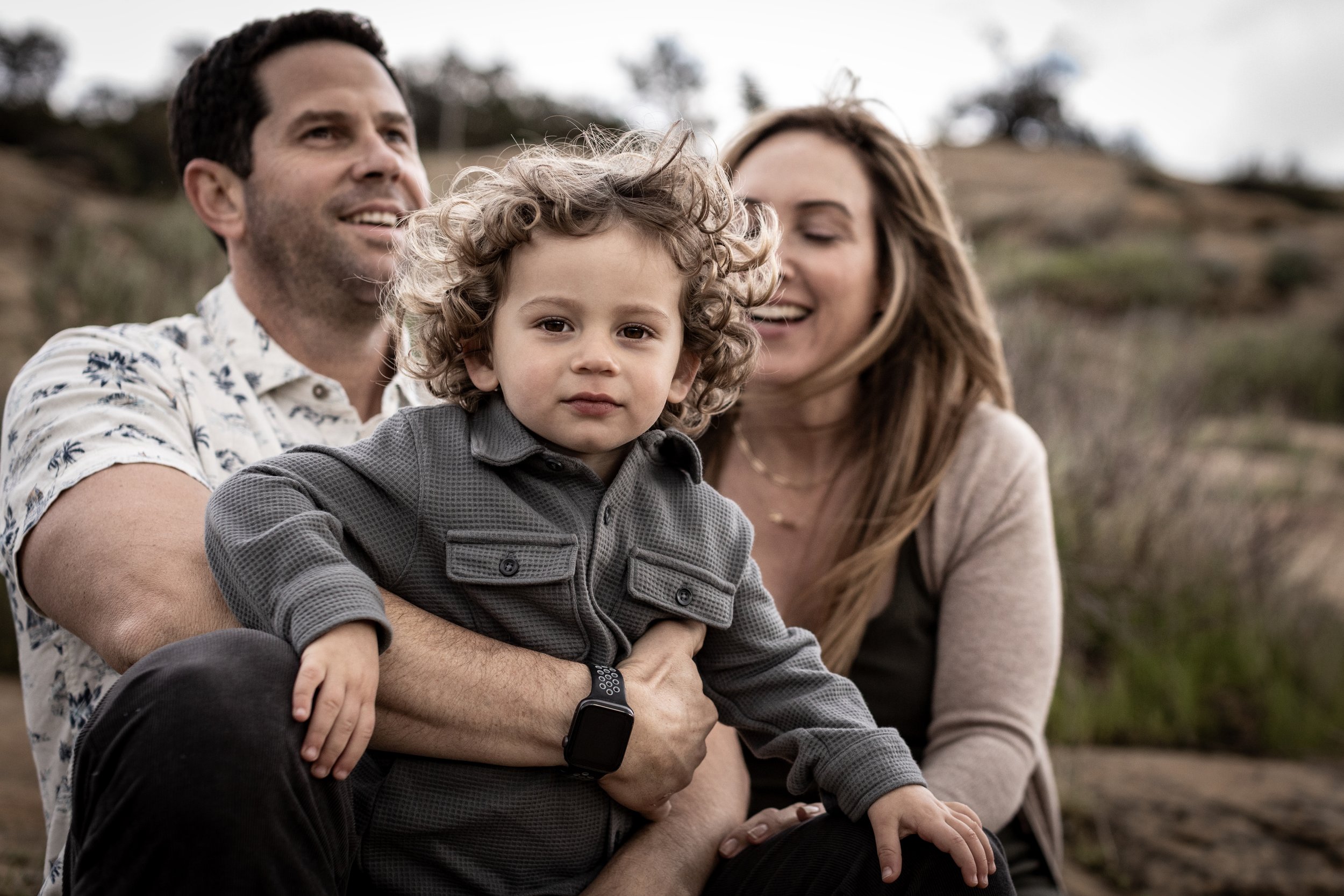 A family of three sitting outdoors on a hillside, with the father holding a young curly-haired boy, and the mother smiling behind them