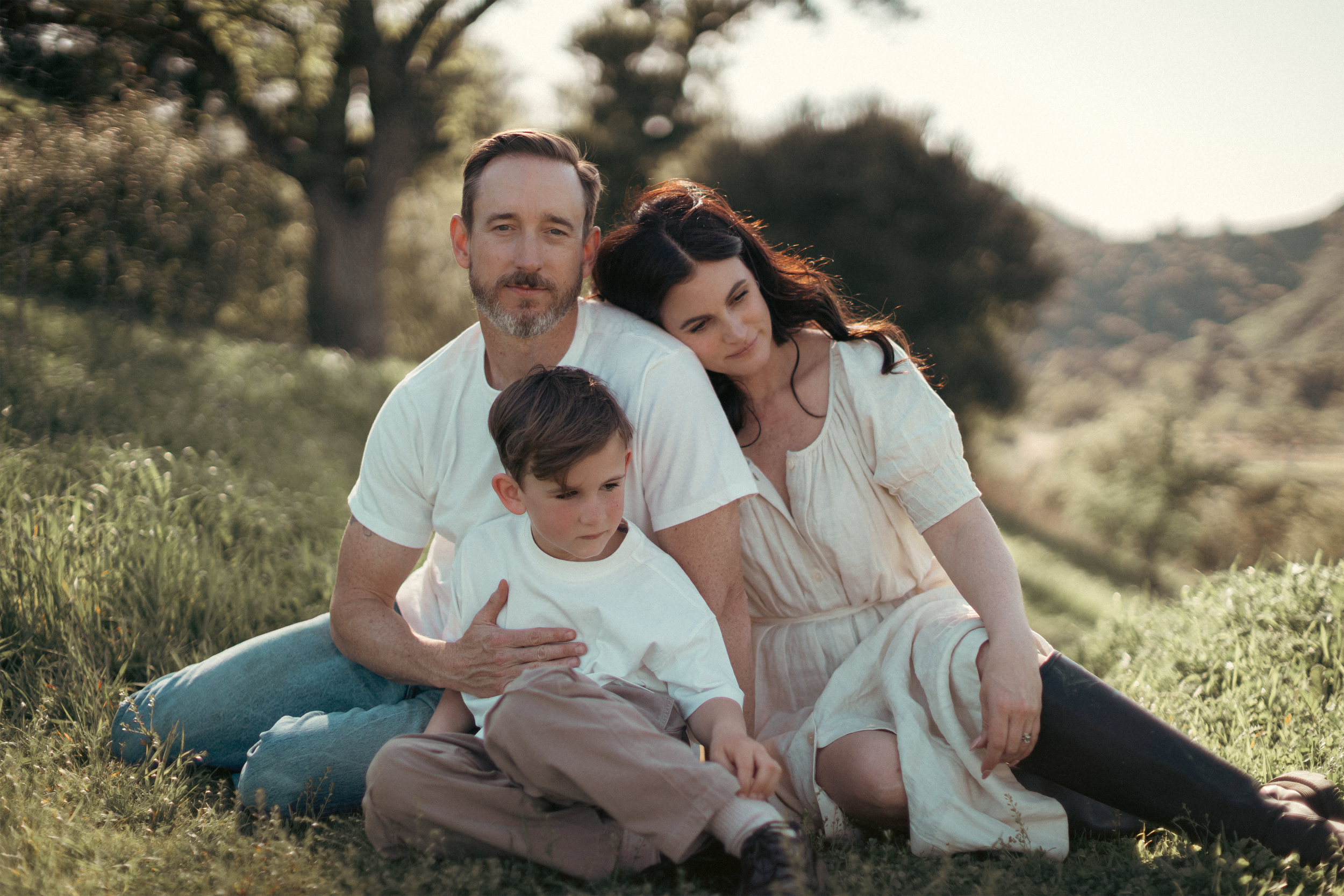 A family of three sitting on grass in a park during sunset, with trees and distant hills in the background.