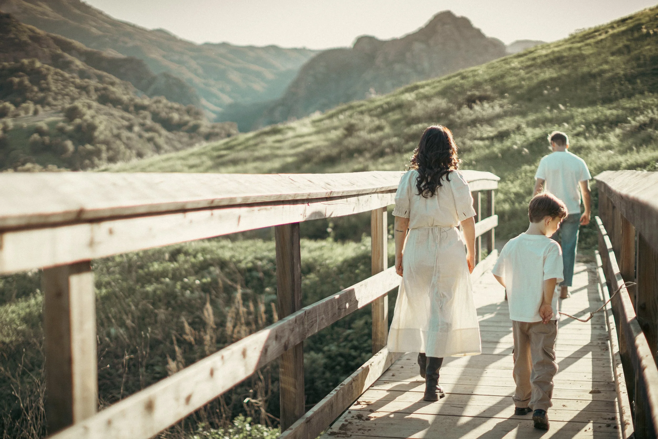 A woman and two children walking along a wooden bridge through a lush, hilly landscape during the daytime.