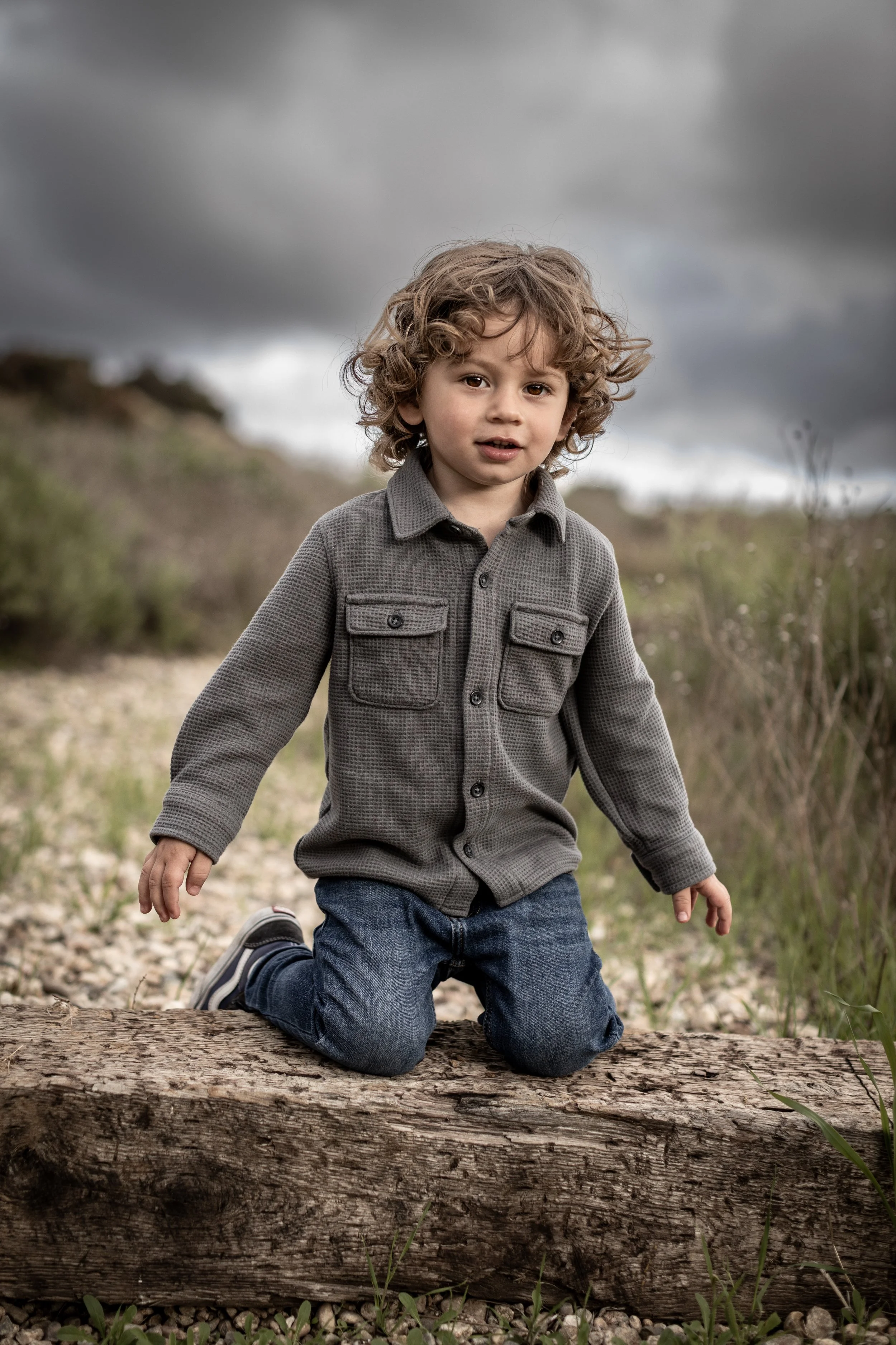 Young boy with curly hair kneeling on a log outdoors on a cloudy day