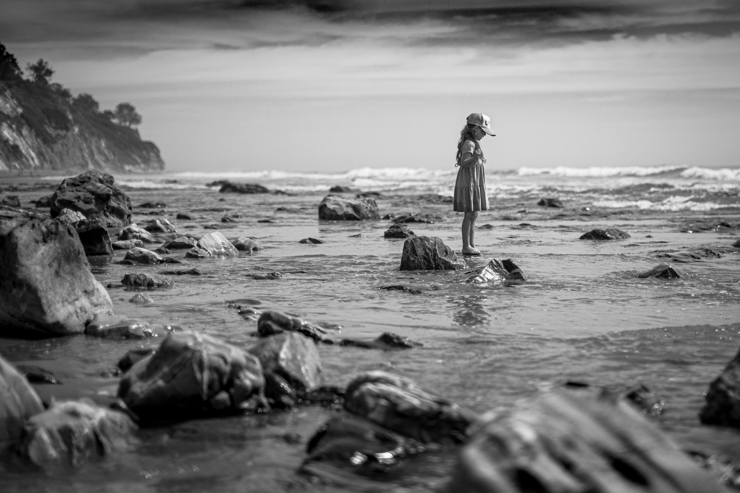 A young girl wearing a dress and a baseball cap stands on rocks in shallow water at the beach, with waves in the background and a cliff on the left side.