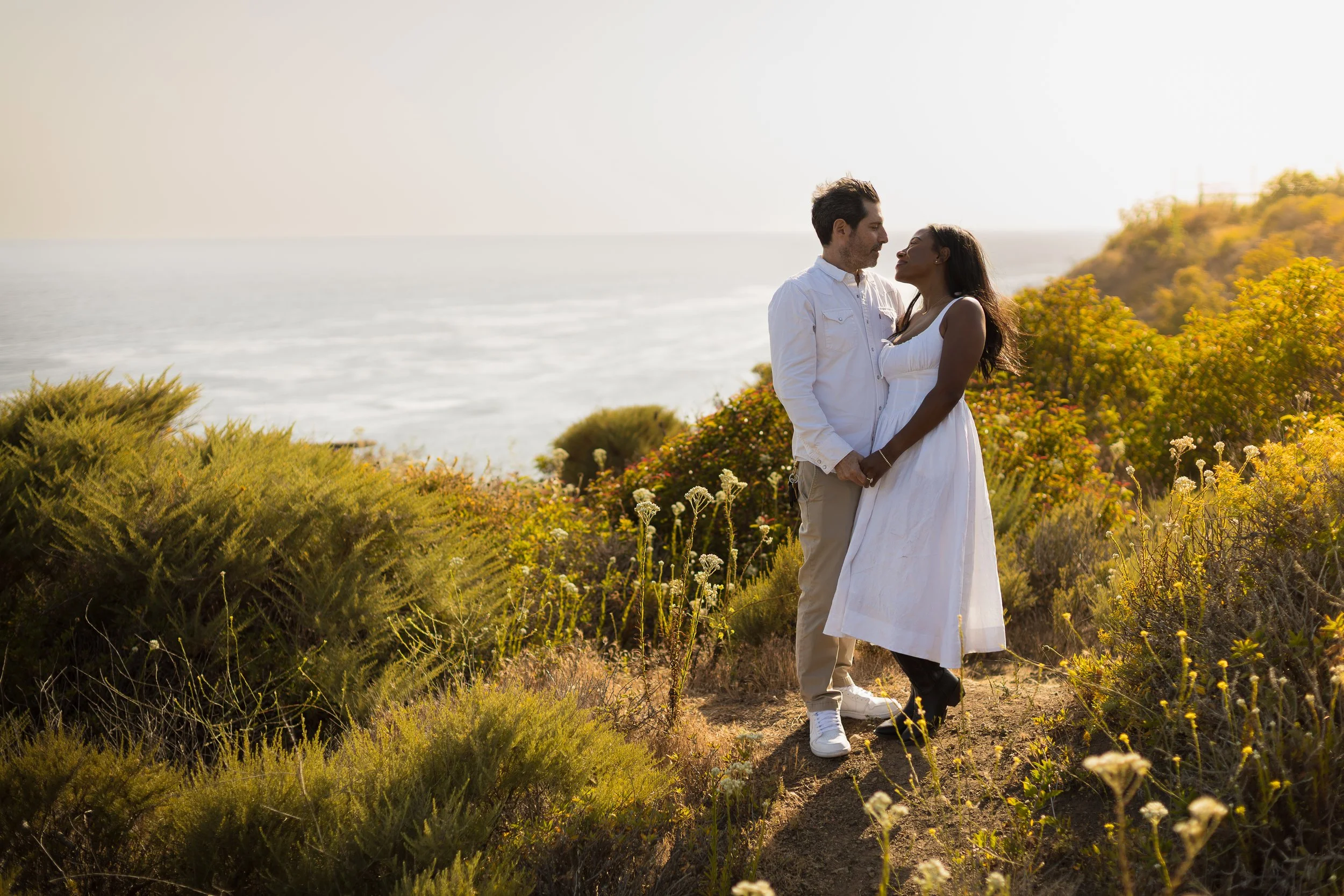 A couple standing hand in hand on a cliffside with coastal vegetation, overlooking the ocean at sunset.