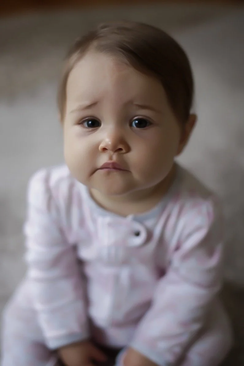 A young child with brown hair and big eyes sitting on the floor, wearing pink and white pajamas, looking up with a slightly sad or worried expression.