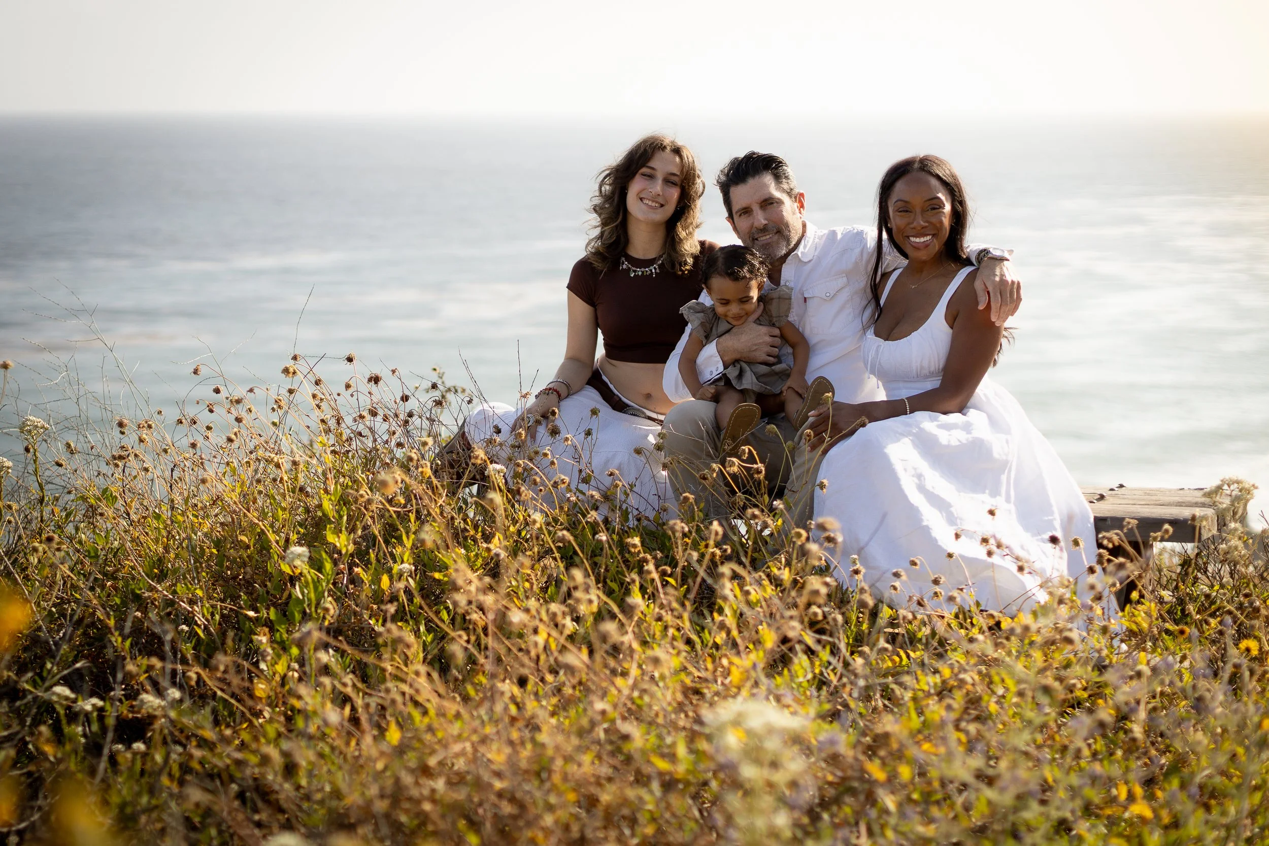 Group of four people, two women, one man, and one child, sitting outdoors near the ocean with tall grasses in front, smiling at the camera on a sunny day.