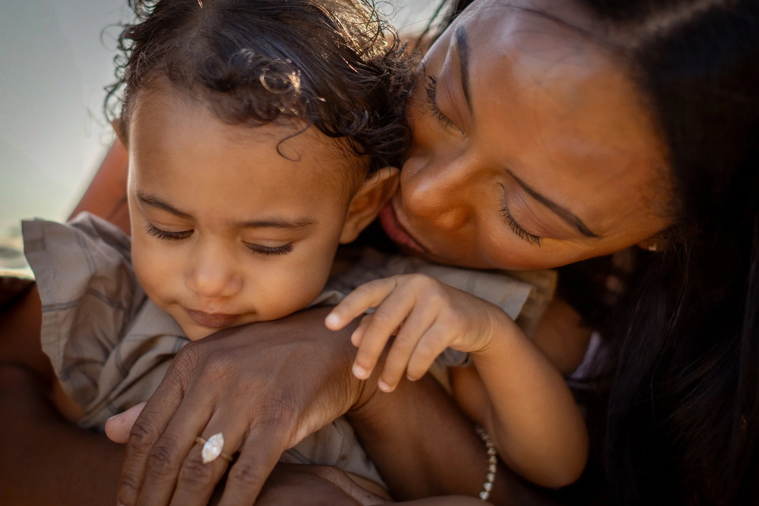 A woman lovingly cuddling a young child, both with their faces close together, eyes closed, appearing to enjoy a tender moment of affection.