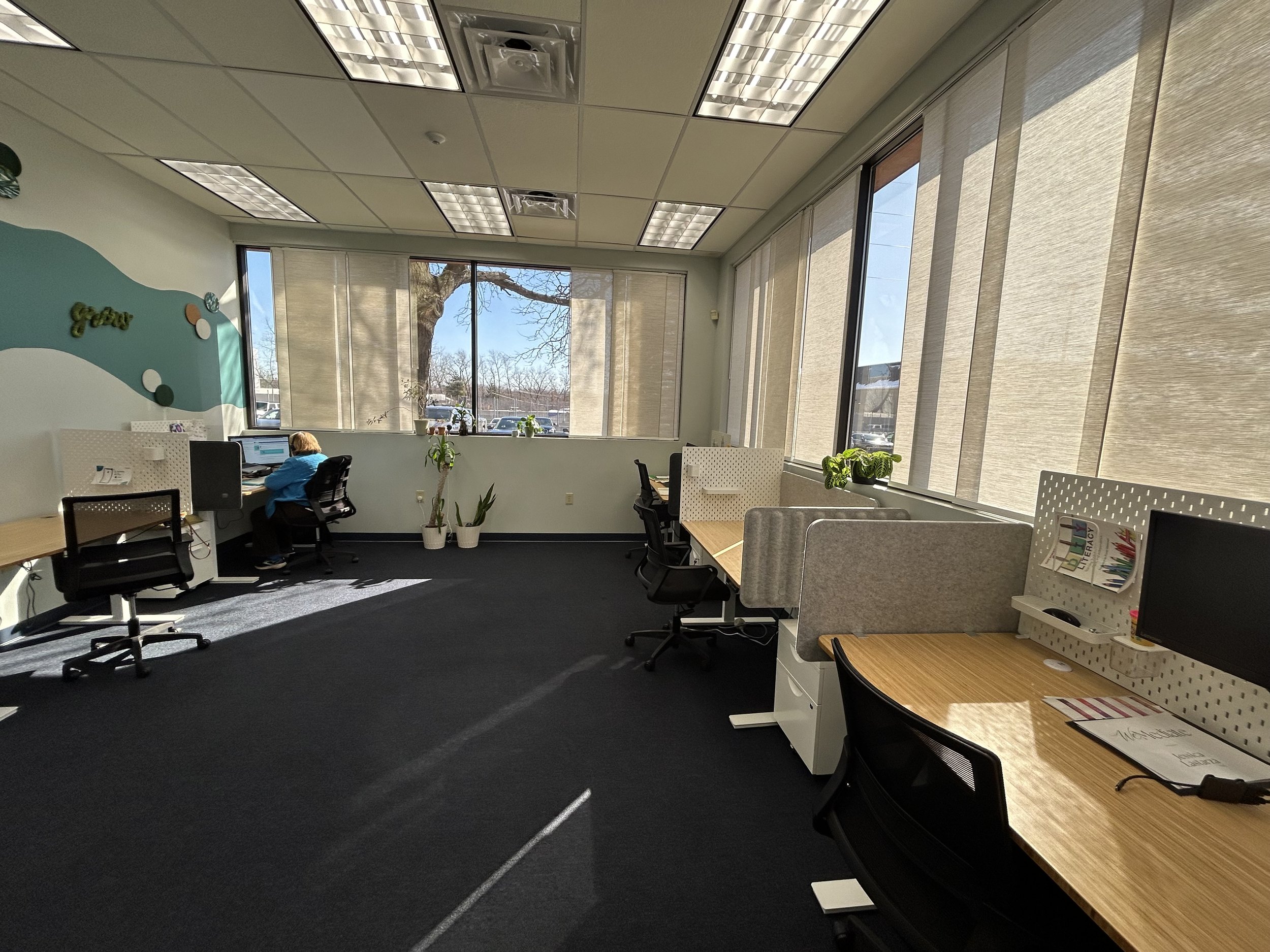 An office room with large windows covered by beige blinds, some of which are partially open, allowing sunlight to stream in and cast shadows on the dark carpeted floor. There are desks with chairs, some occupied by individuals, and various plants are placed on the windowsills and desks. The ceiling has recessed lighting fixtures.