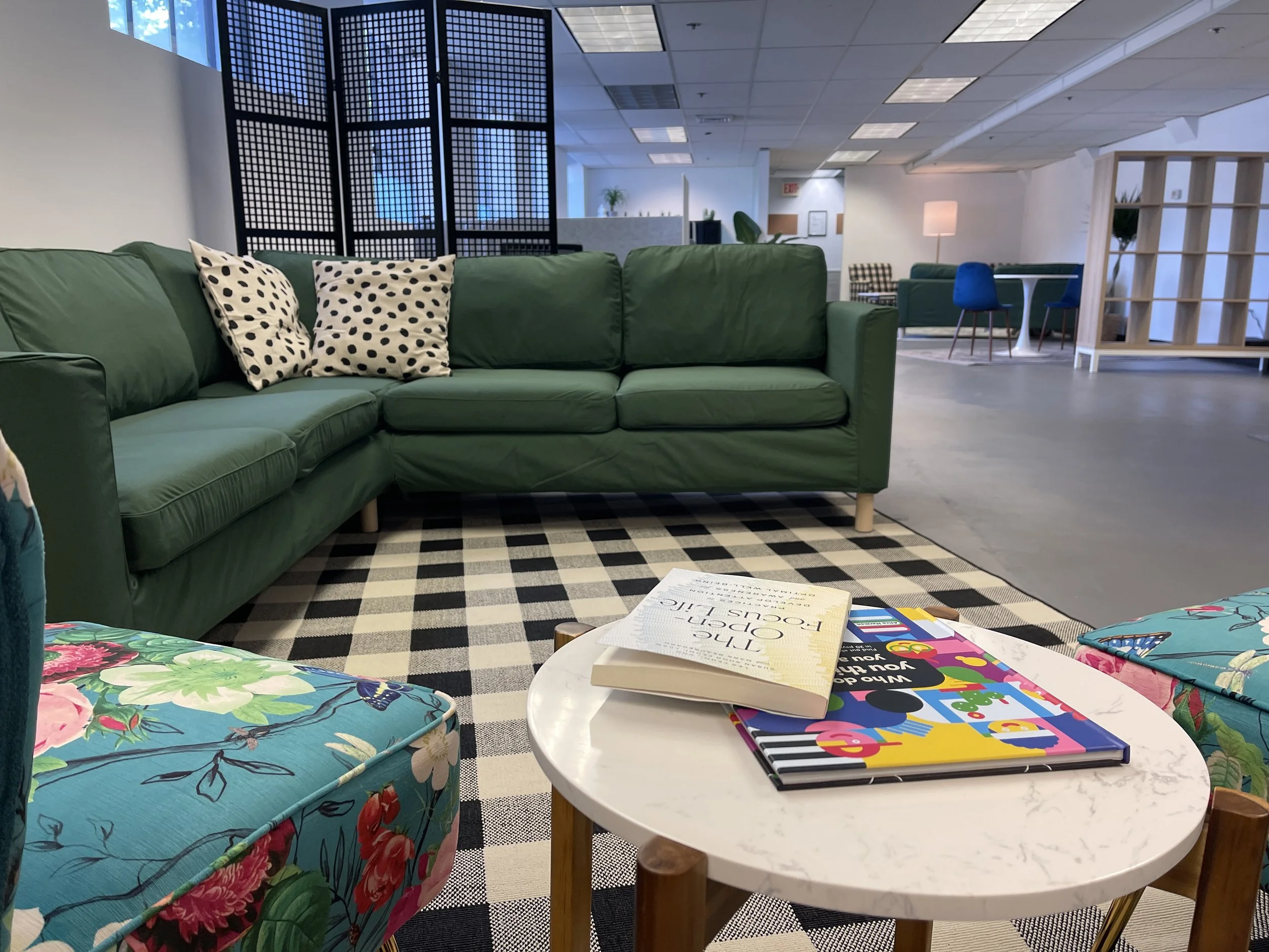 A modern lobby area with green sofa and decorative pillows, black and white checkered rug, white round table with books, colorful floral upholstered chairs, a wooden room divider, and other seating with a high ceiling and fluorescent lighting.