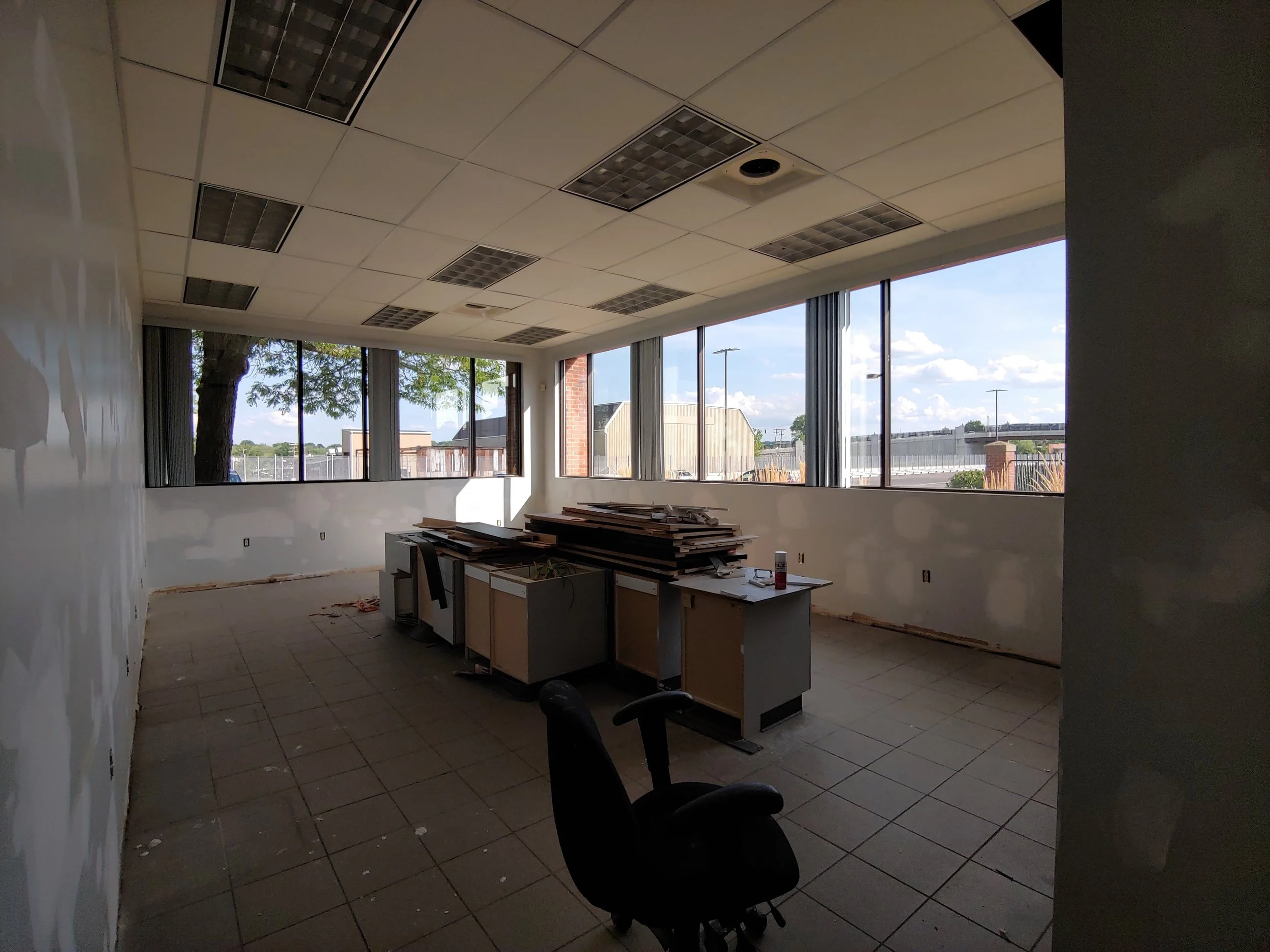 An empty office room under construction or renovation with large windows, ceiling lights, stacks of wood, and a black office chair. Farese & Co. Strategic Commercial Space Strategy for Property Owners and Lessees West Hartford Coworking