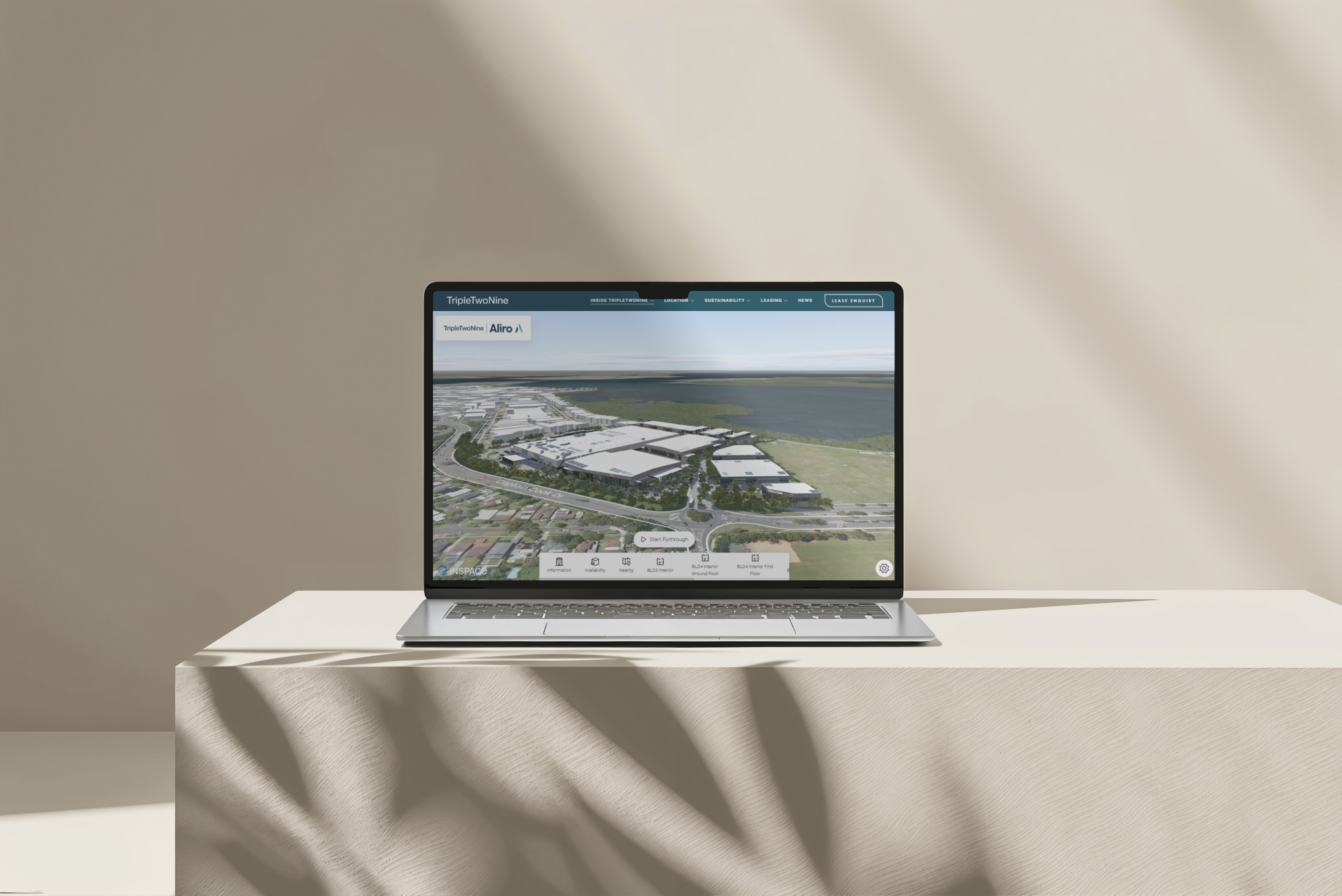 Laptop on a light-colored table displaying an aerial view of a modern urban development project, overlooking a body of water, with shadows cast on the table.