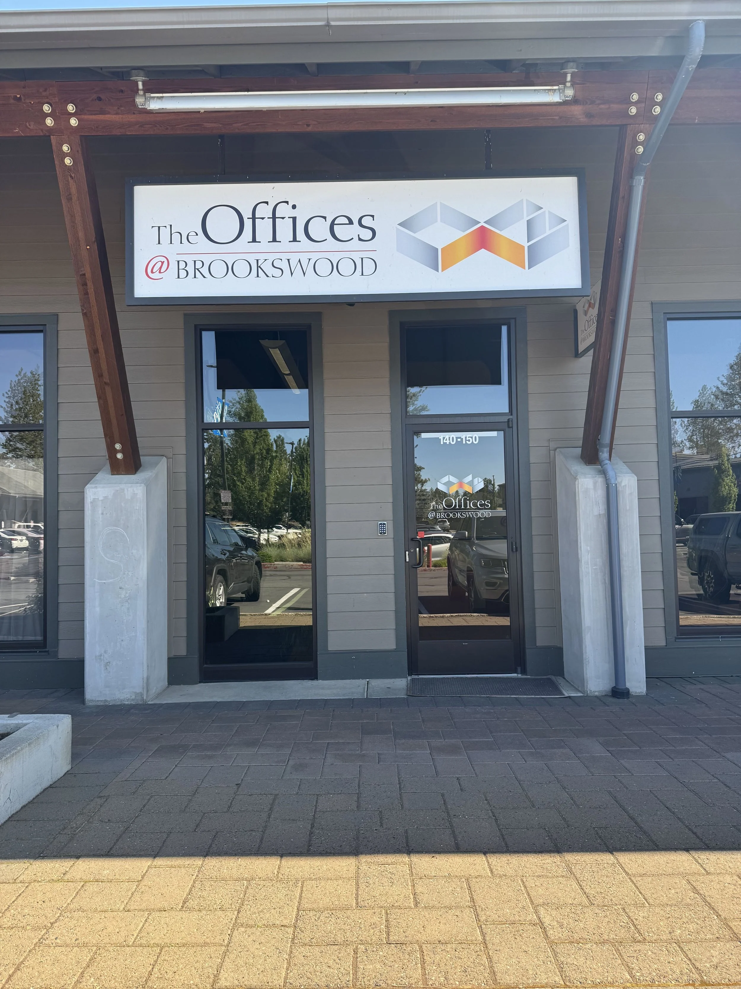 Storefront with a sign that reads 'The Offices @ Brookwood,' featuring a geometric logo and windows reflecting a parking lot and trees.