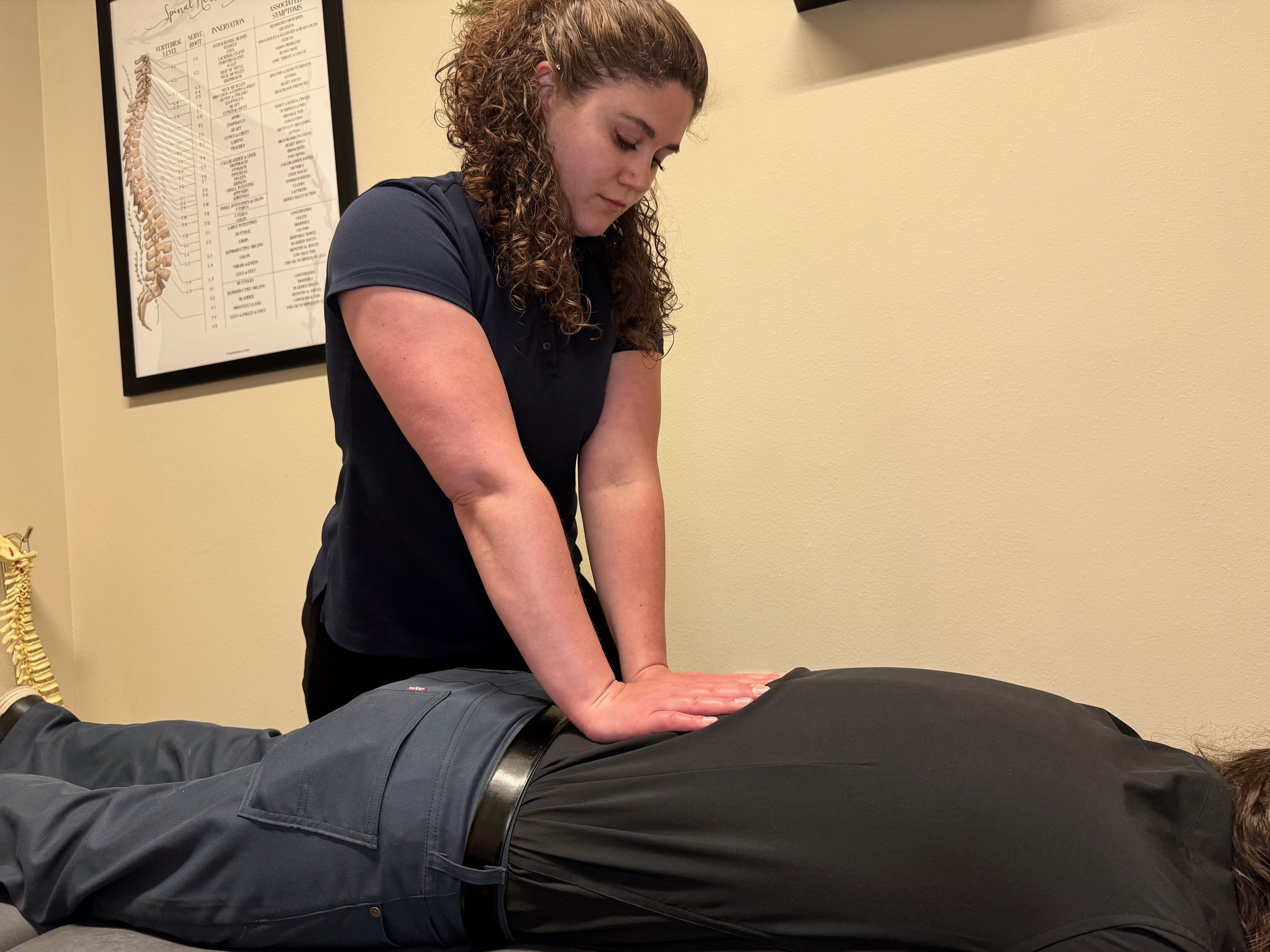 A woman with curly hair giving a massage to a person lying face down on a table in a therapy room with a spine chart on the wall.