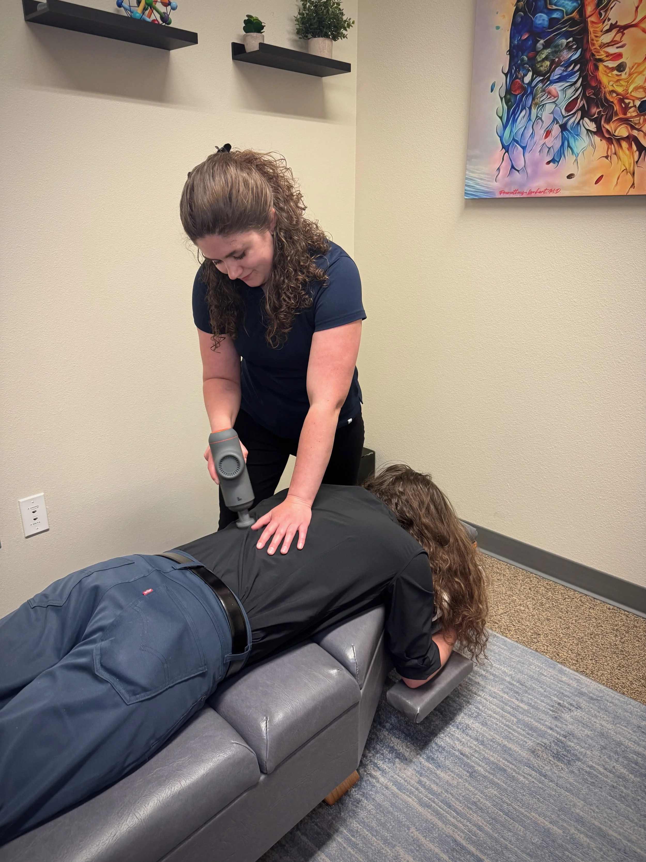A woman receiving physical therapy, using a massage gun on a man's back while he lies face down on a sofa in an indoor setting.
