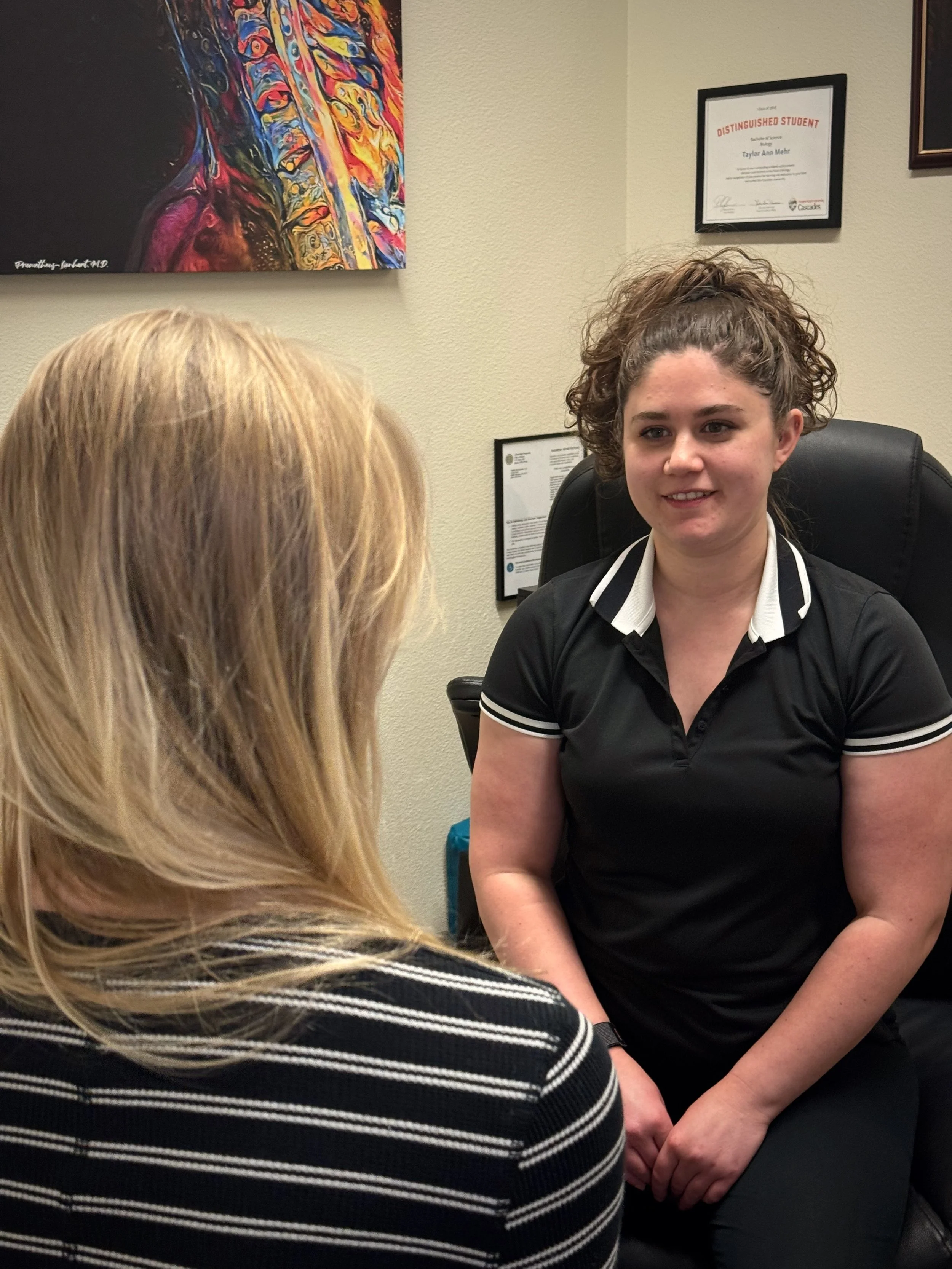 Two women are sitting and talking, one with curly brown hair wearing a black shirt with white trim, and the other with blonde hair wearing a black and white striped shirt. There is a colorful painting on the wall and a framed certificate in the background.