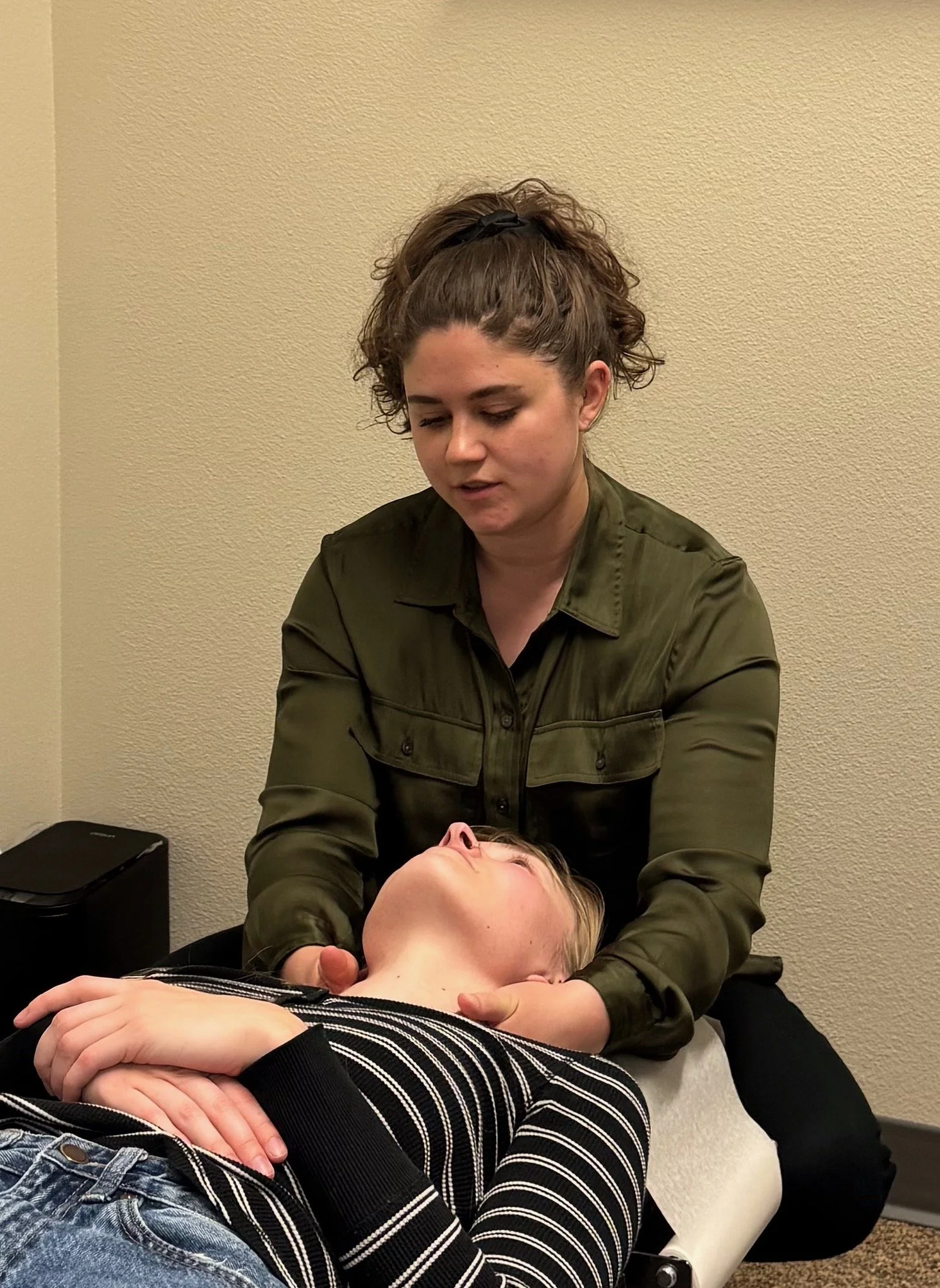 A person with curly hair and a black hair scrunchie is practicing chiropractic or massage therapy on a woman lying face-up. The woman on the table has blonde hair and is wearing a striped long-sleeve shirt and jeans.