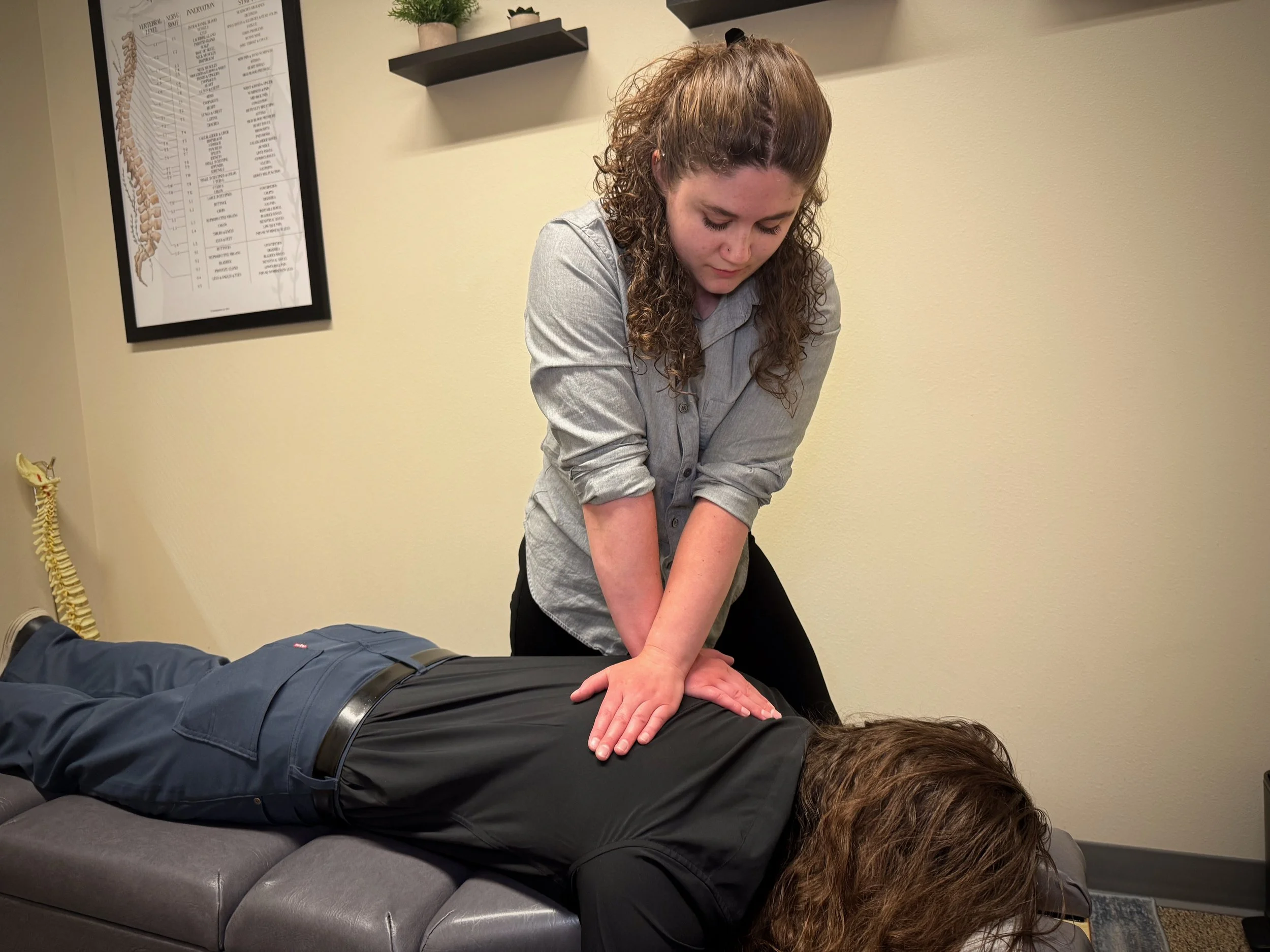 A woman performing CPR on a man lying face down on a padded surface, with a chiropractor or medical professional supervising in a clinical setting.