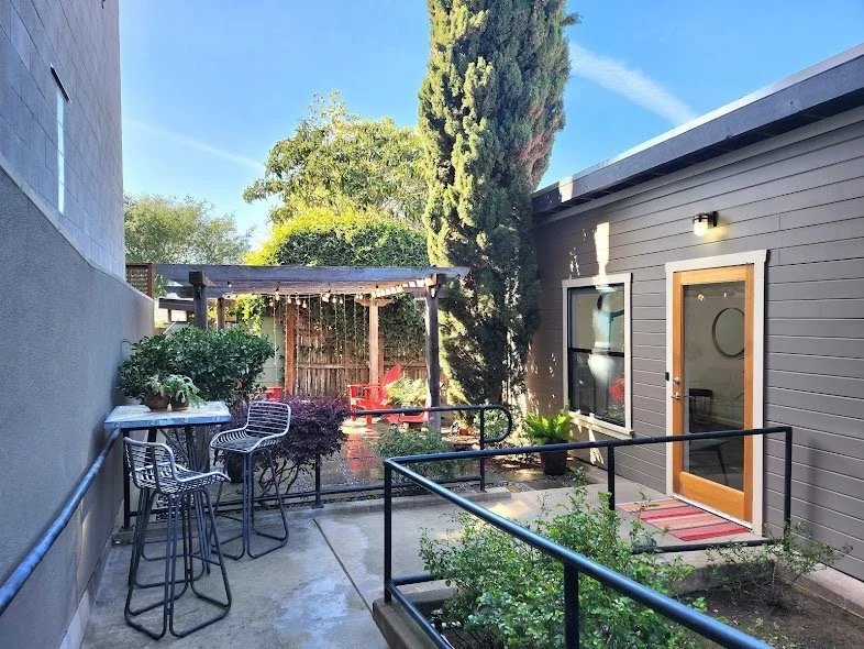 Outdoor patio area with seating and greenery, adjacent to a modern gray building with a glass door and a window, under a clear blue sky.
