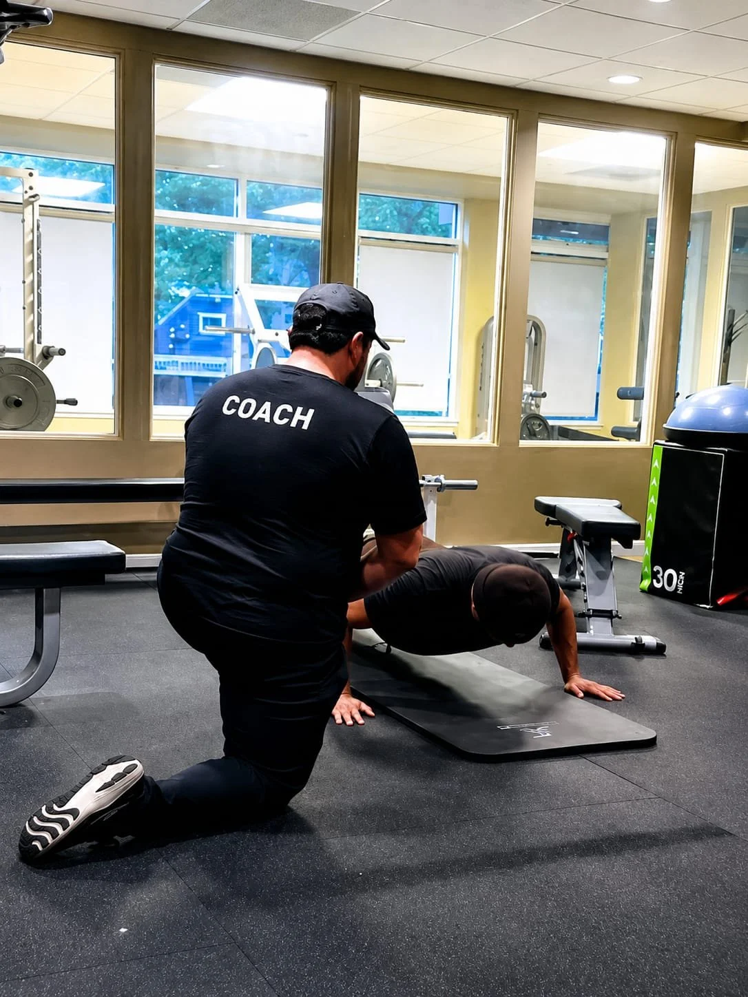 A personal trainer wearing a black shirt labeled 'COACH' assists a person performing a plank exercise on a mat in a gym with large windows.
