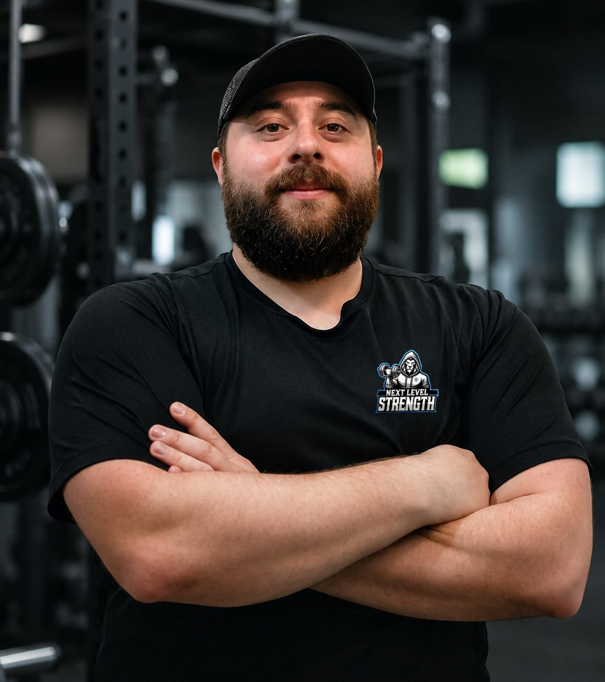 A man with a beard and mustache wearing a black t-shirt with the logo 'Next Level Strength' and a black cap, standing with arms crossed in a gym with weightlifting equipment in the background.