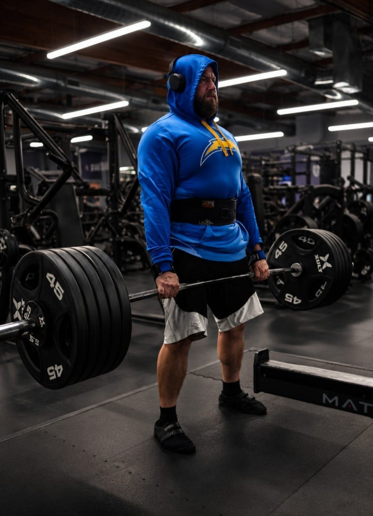 A man lifting a heavy barbell with black weights at a gym. He is wearing a blue hoodie with a yellow lightning bolt logo, headphones, black shorts, and black sandals.