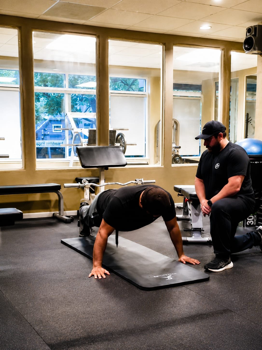 A man doing push-ups on a yoga mat in a gym with a trainer kneeling beside him. The gym has large windows, exercise equipment, and a black rubber floor.