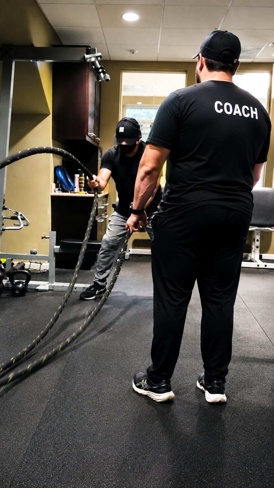 A personal trainer wearing a shirt labeled 'COACH' is instructing a client performing a battle rope exercise in a gym.