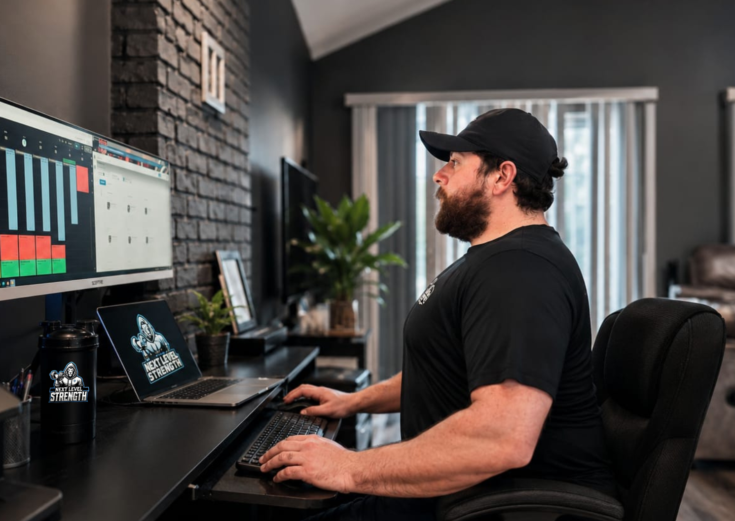 A man with a beard and wearing a black cap and shirt sitting at a desk working on a computer. On the desk are a laptop, a water bottle, and a monitor displaying a bar graph and a calendar. The room has gray walls, a large window with curtains, and in the background, there are plants and picture frames.