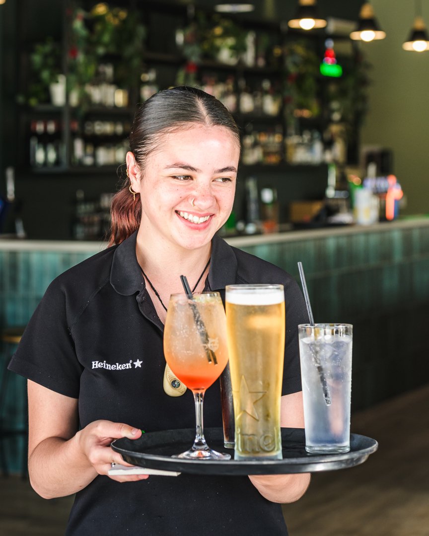 A smiling server holds a tray with three drinks: a cocktail, a pint of beer, and a glass of water. The bar setting is visible in the background.