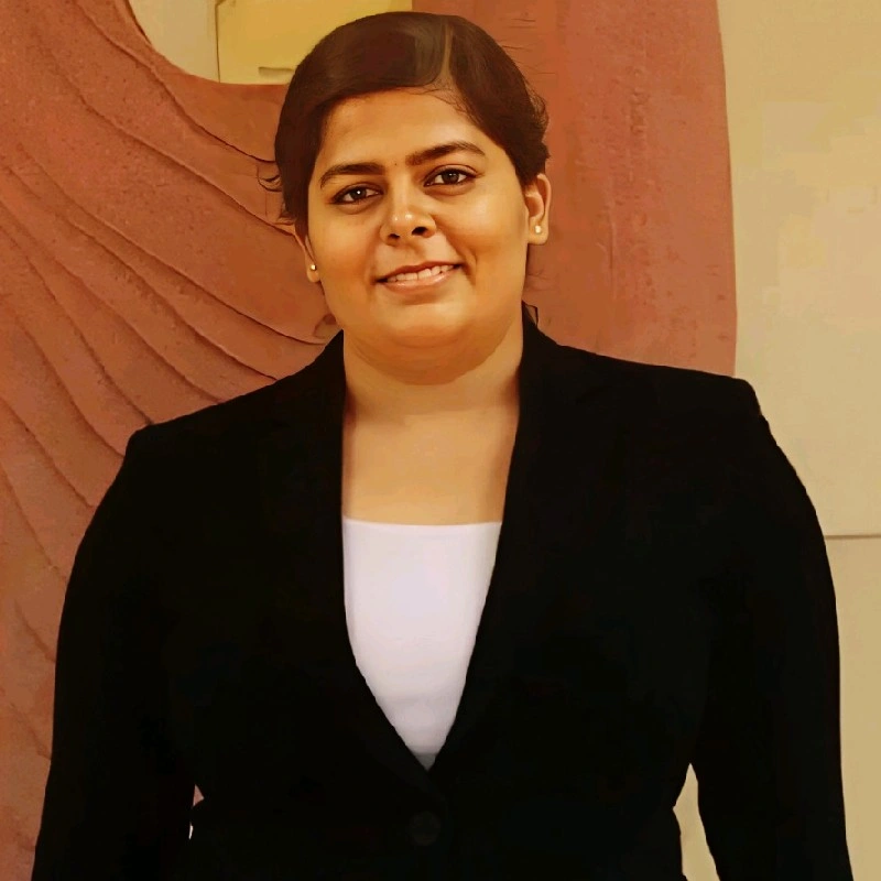 A woman with short dark hair, wearing a black blazer over a white top, smiling in front of a textured, curved wall.