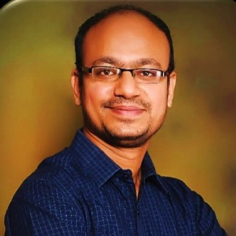 A man with glasses and a beard, smiling, wearing a dark blue shirt, against a blurred green and brown background.
